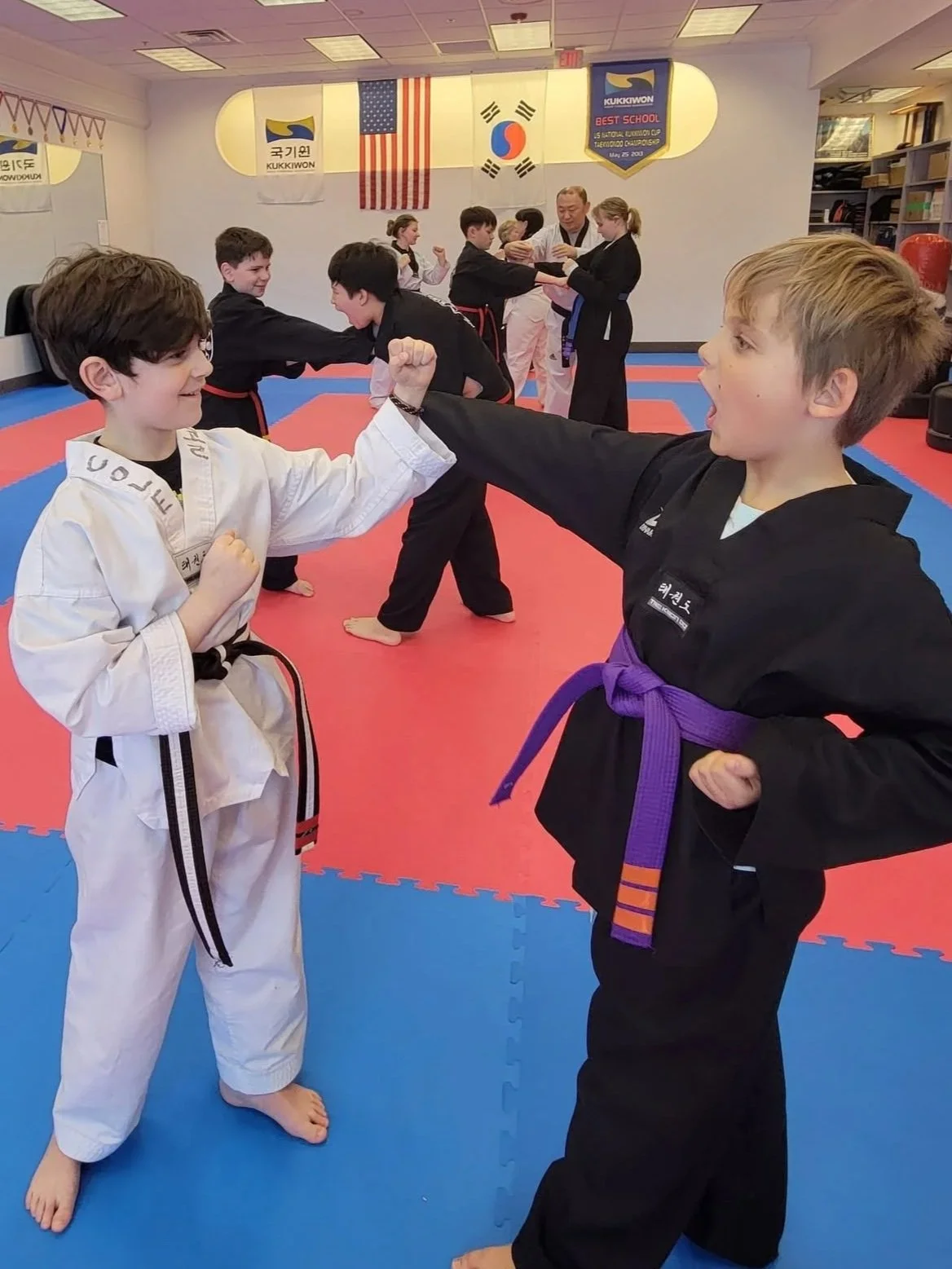 Two children in taekwondo uniforms sparring in a taekwondo dojang with banners and flags on the wall, while other students practice in the background.
