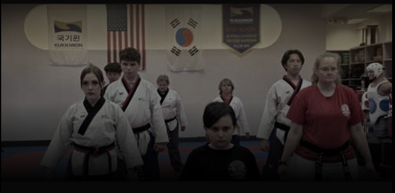 Group of students in a taekwondo dojang, wearing white doboks and belts, with flags of South Korea, the United States, and South Korea banners hanging on the wall.