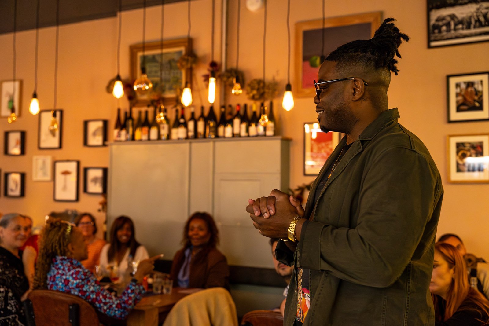 A man with glasses and dreadlocks tied up, wearing a dark jacket and gold watch, is standing in a restaurant or bar, smiling with his hands clasped. In the background, people are seated at tables, laughing and engaging, with warm lighting and framed pictures on the wall.