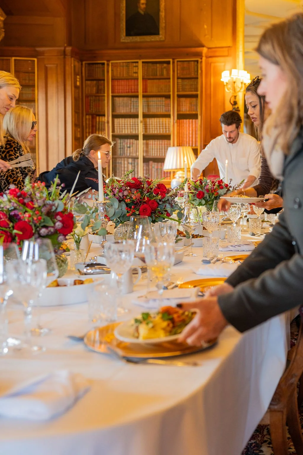 People gathered around a elegantly set dining table with floral centerpieces, candles, and fine glassware in a wood-paneled room.