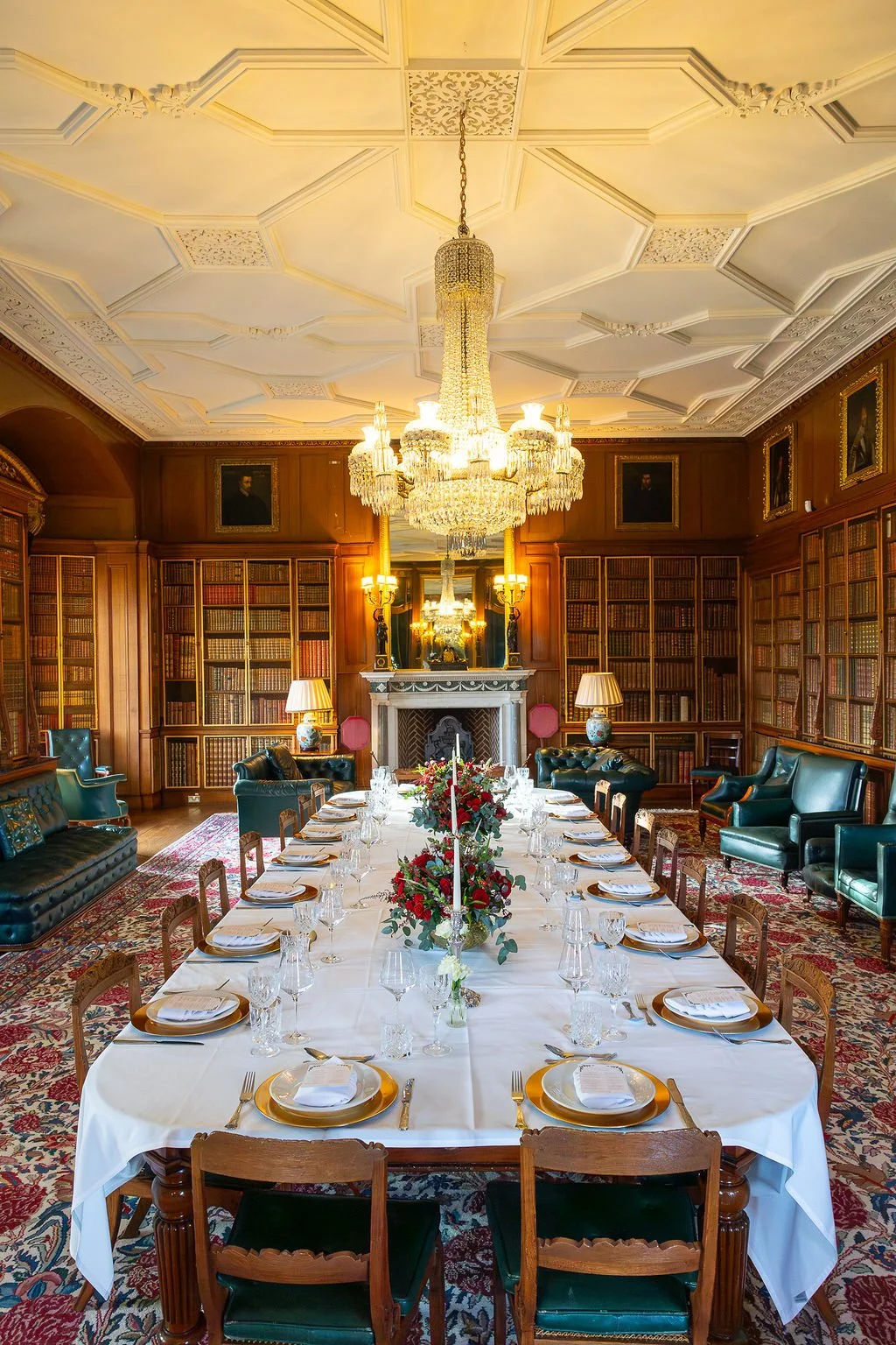 Elegant dining room with a long table set for a formal meal, surrounded by wooden chairs, and decorated with floral arrangements. The room features wood-paneled walls, built-in bookshelves, a marble fireplace, and a large ornate chandelier hanging from the decorated ceiling, with framed portraits on the walls.