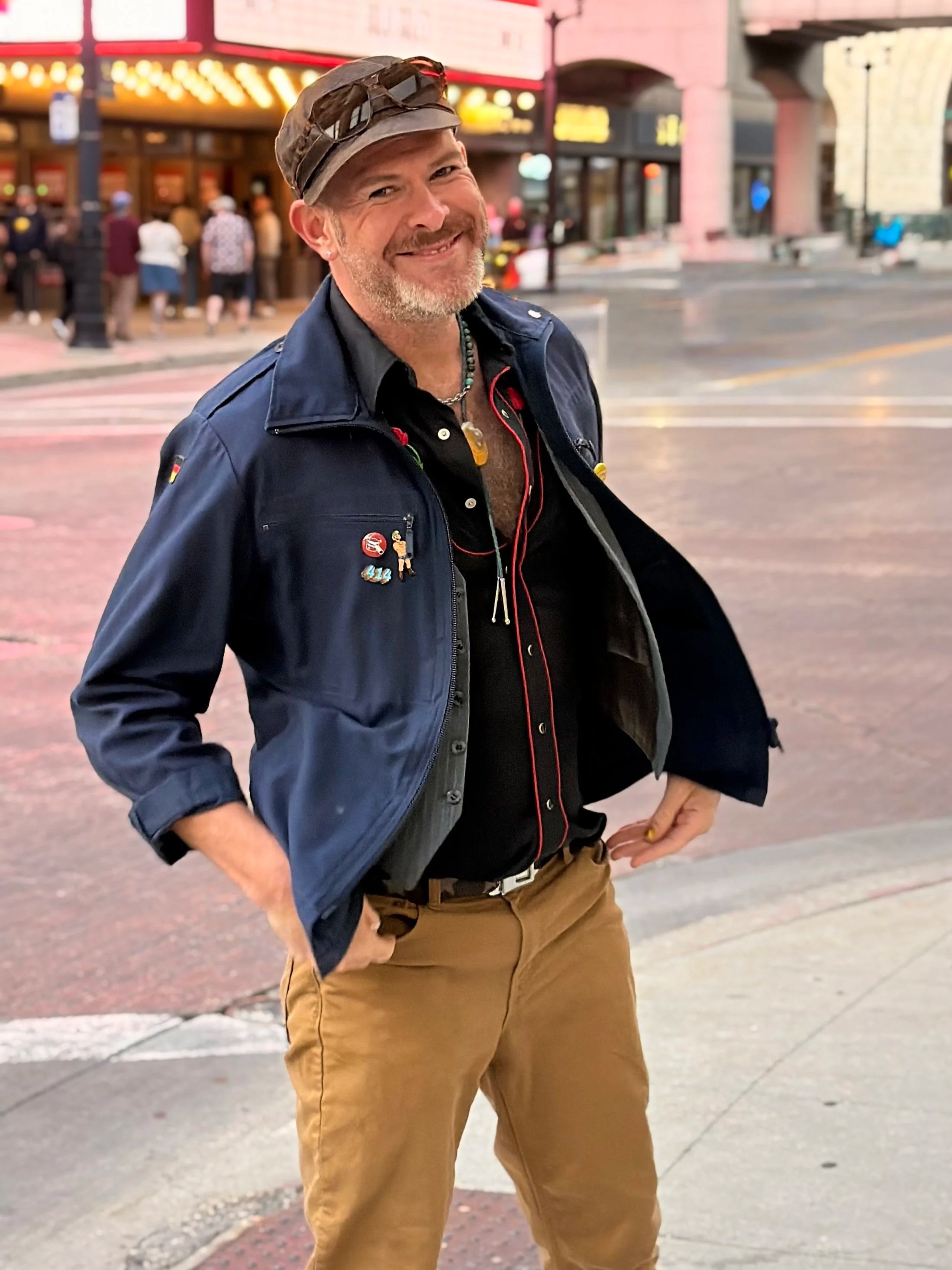 A smiling man wearing a dark blue jacket, khaki pants, a baseball cap, sunglasses on his hat, and a necklace, standing on a city sidewalk with a busy street, colorful buildings, and pedestrians in the background.