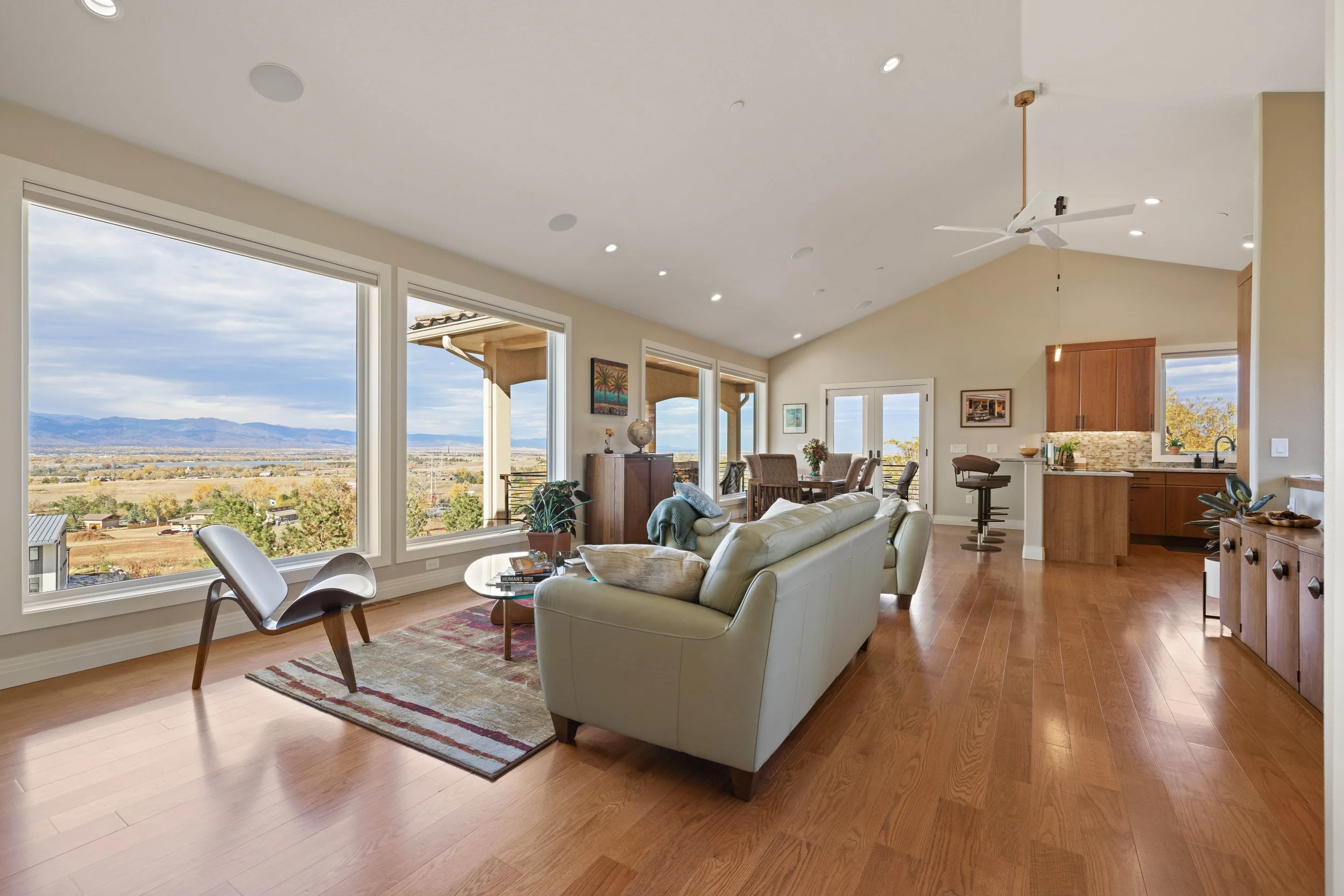 Lucky Lopez's real estate photography - Living room with large windows showing a mountainous landscape, light-colored sofa, modern chair, wooden furniture, ceiling fan, and hardwood floors.