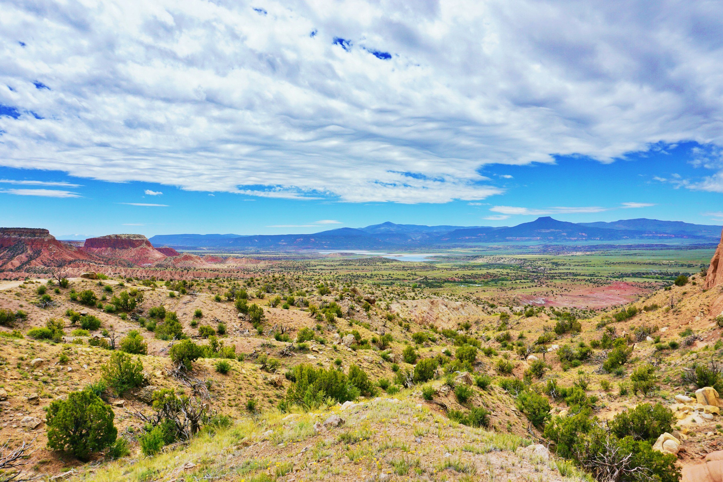 A expansive desert landscape with sparse green shrubbery, rugged red rock formations, and distant mountains under a partly cloudy sky.