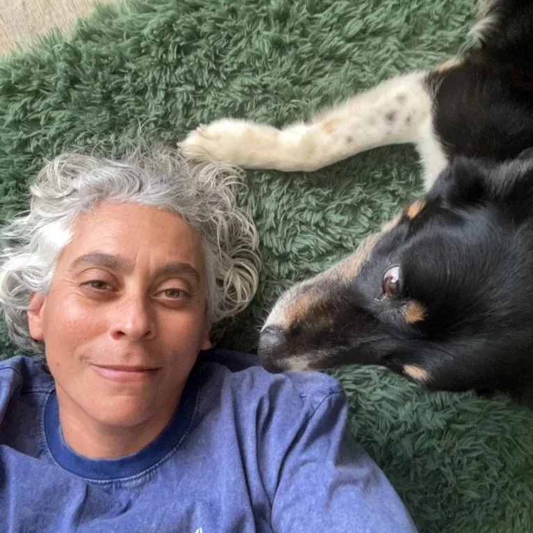 A woman with curly gray hair lying on a green textured rug next to a black and tan dog with a white chest, both looking towards each other.
