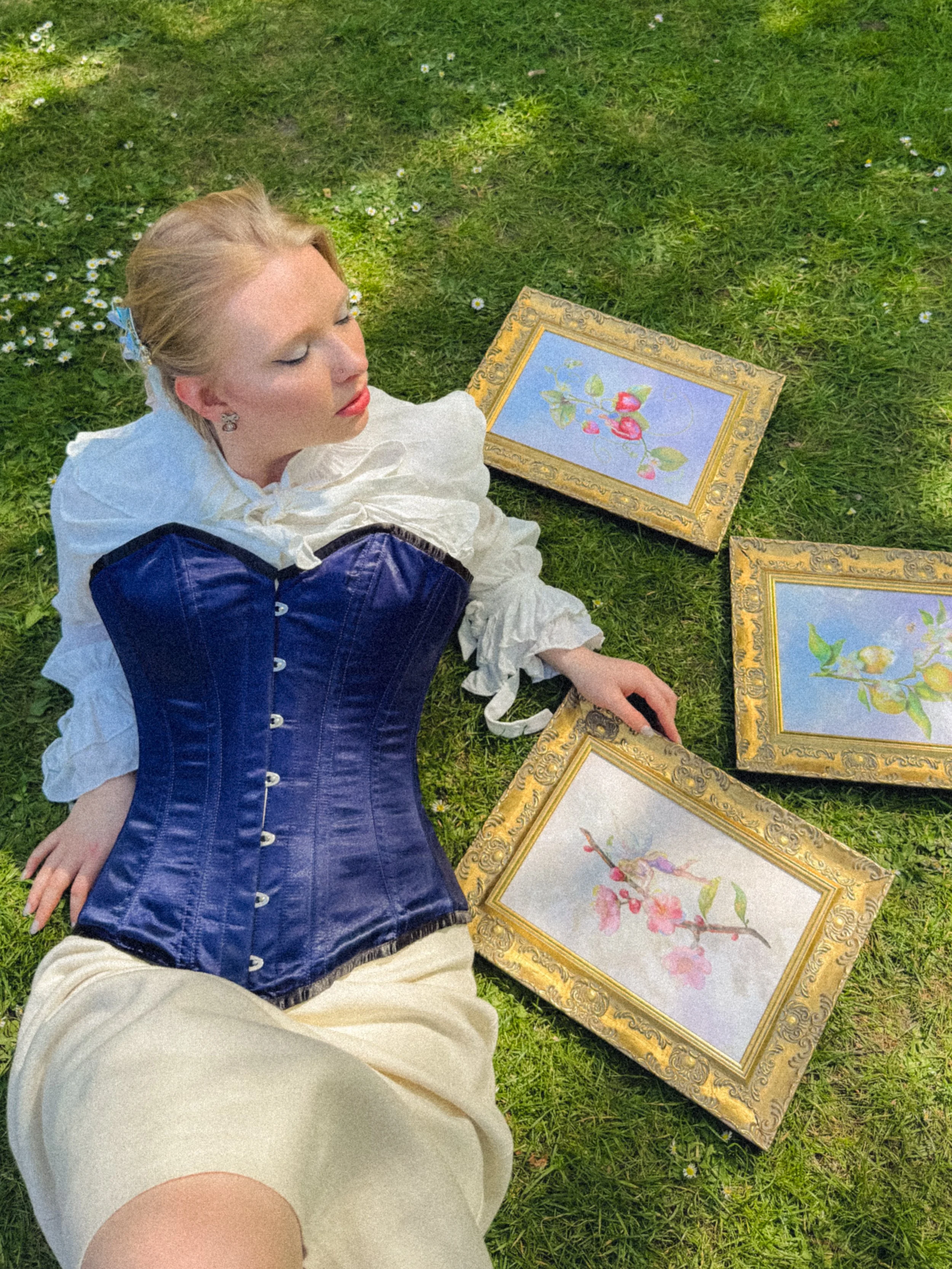 A woman with blonde hair, dressed in a white and blue outfit, is lying on green grass surrounded by floral paintings with ornate gold frames. The paintings depict pink flowers and green foliage.