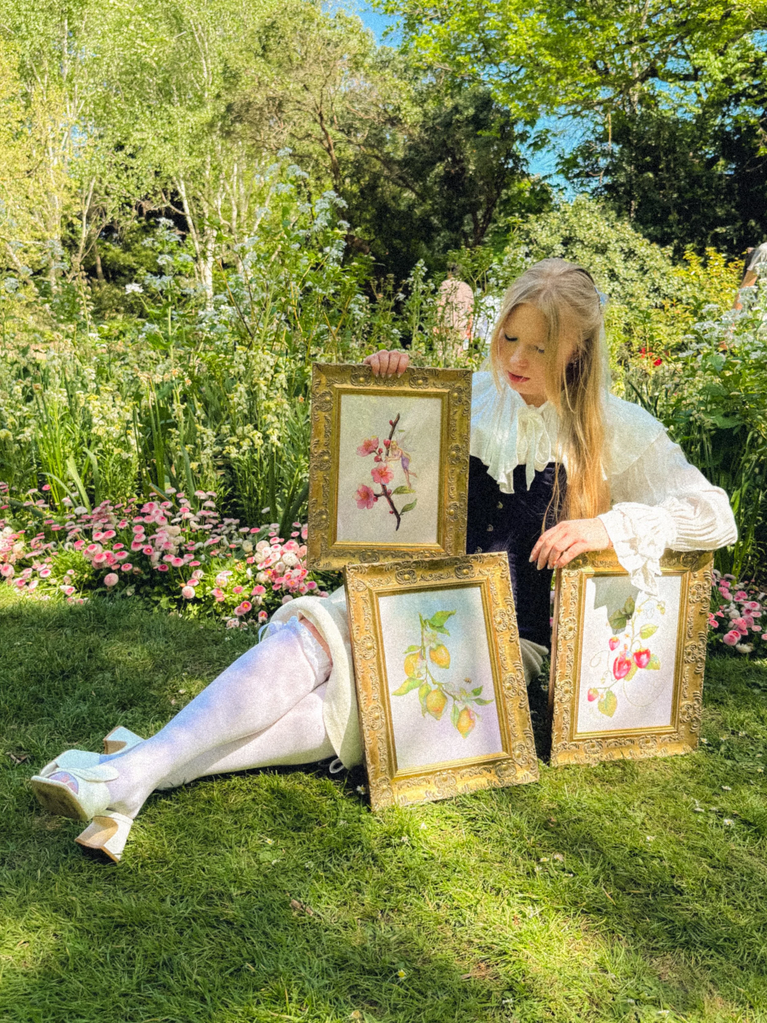 A woman sitting on the grass in a lush garden surrounded by flowers, holding framed botanical illustrations of fruit trees.