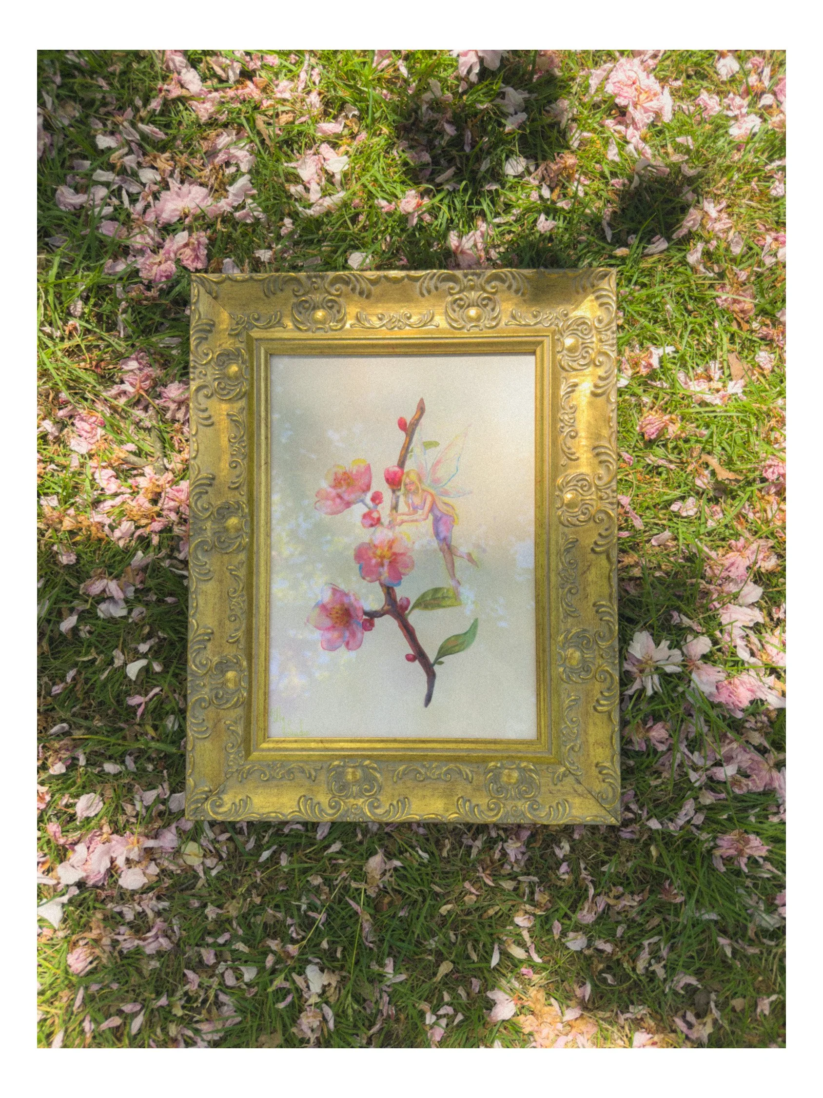 A decorative framed picture lying on grass with pink flower petals scattered around, and a small fairy illustration on the picture's canvas.