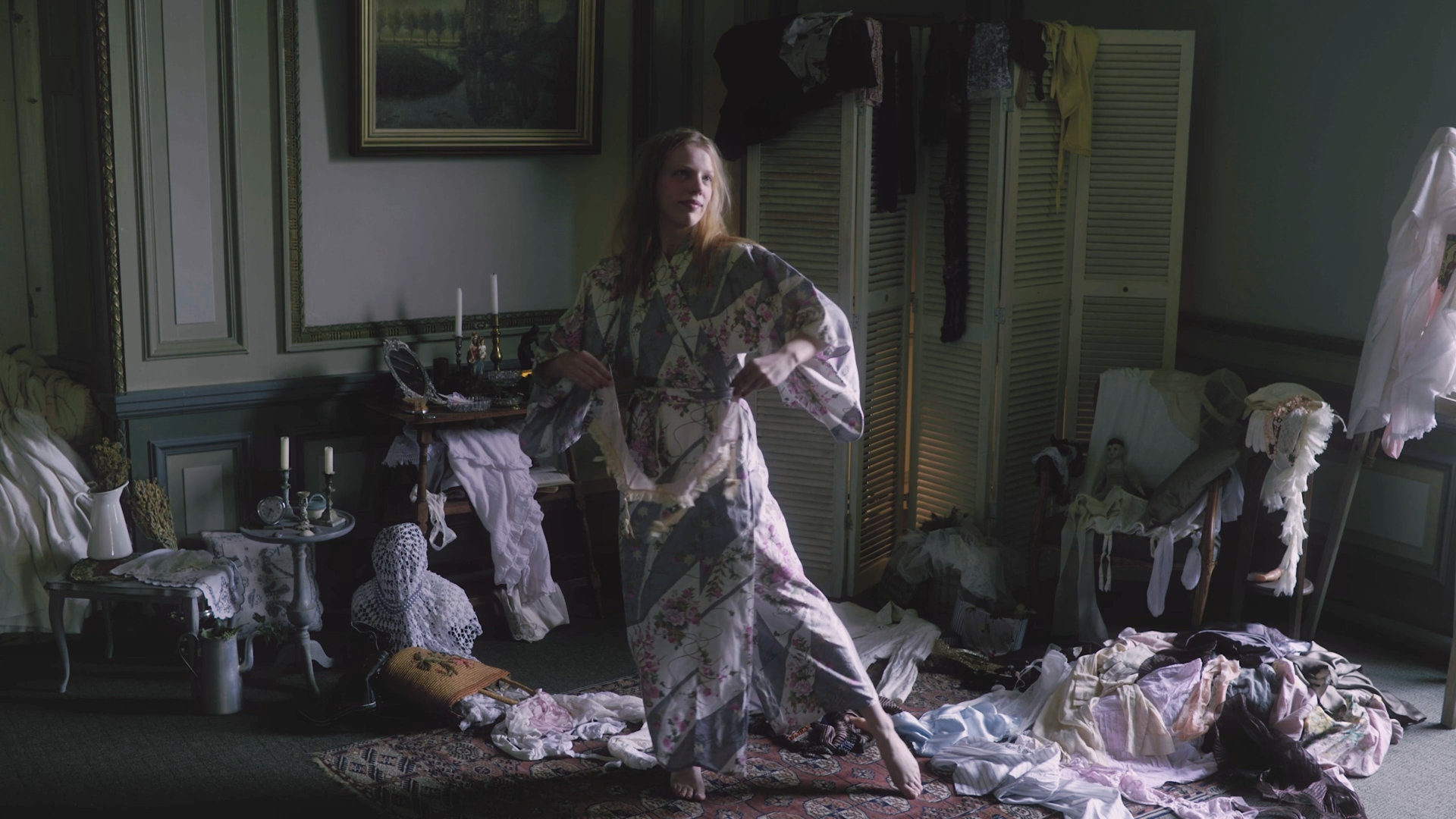 A woman in floral pajamas standing in a cluttered living room, surrounded by laundry, clothes, and decorative items, with a dark green wall and window in the background.