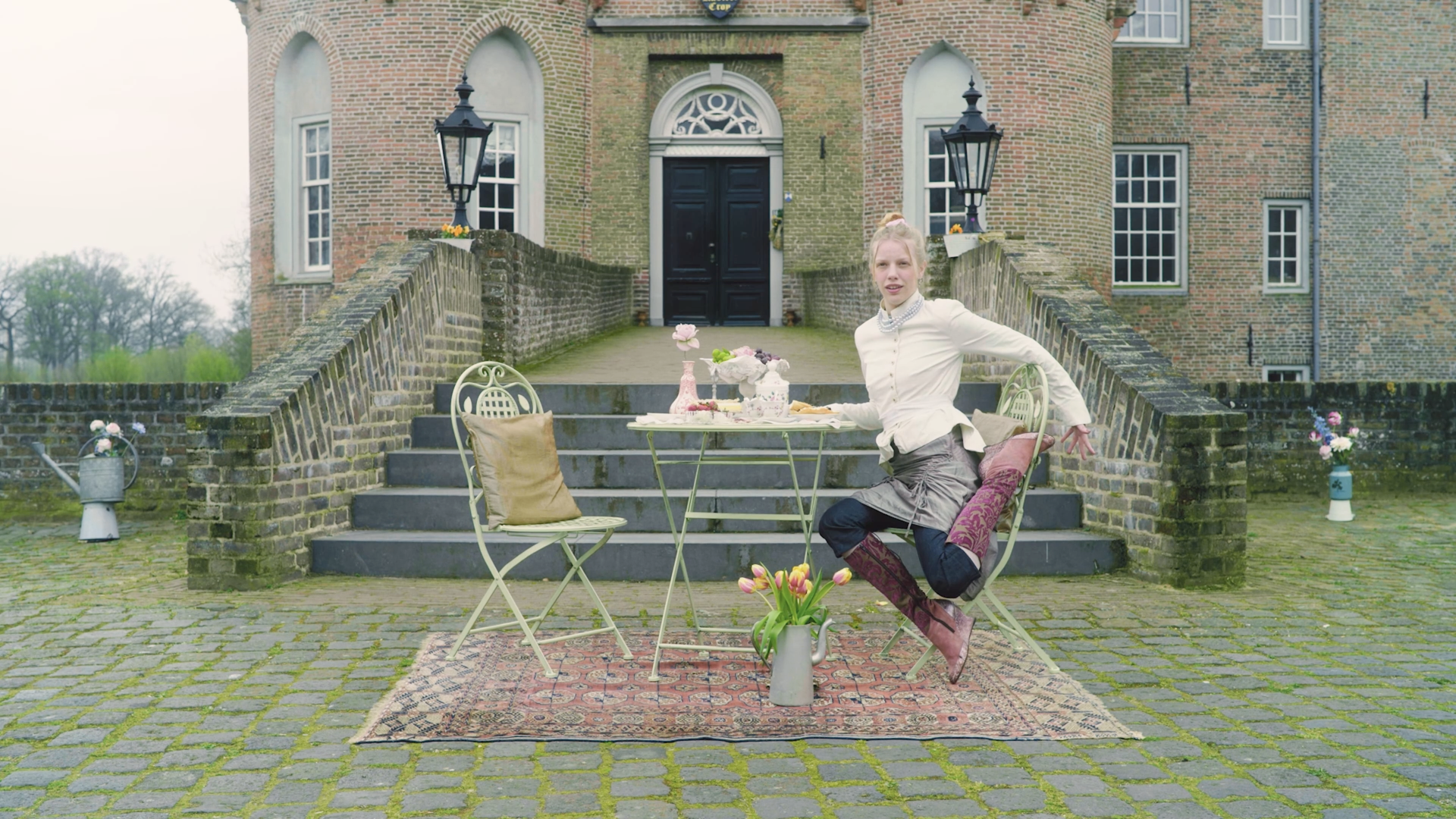 A woman sitting at an outdoor table set for tea or coffee, with a large brick and stone house in the background. The table has a pink vase with flowers, pastries, and teacups. The woman is dressed in vintage clothing, with a light shirt, dark skirt, and patterned stockings, and she is adjusting her position on the chair.