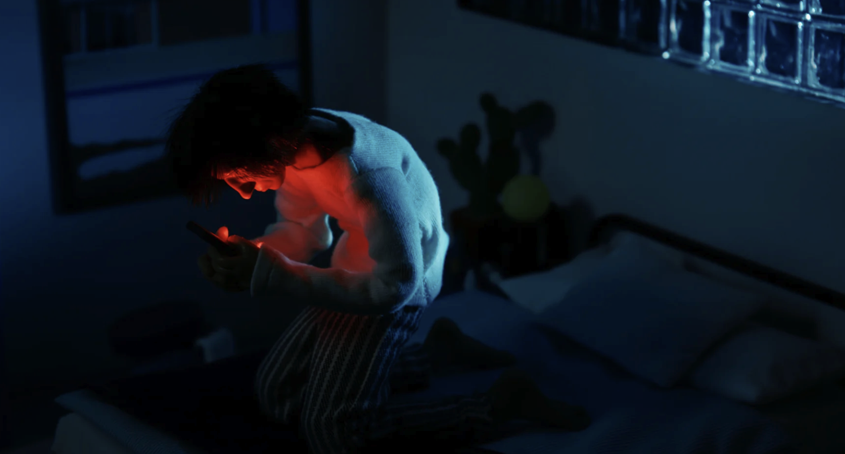 Person sitting on bed at night, illuminated by red light from their phone, in a dimly lit room with glass blocks on the window and a small plant on a table.