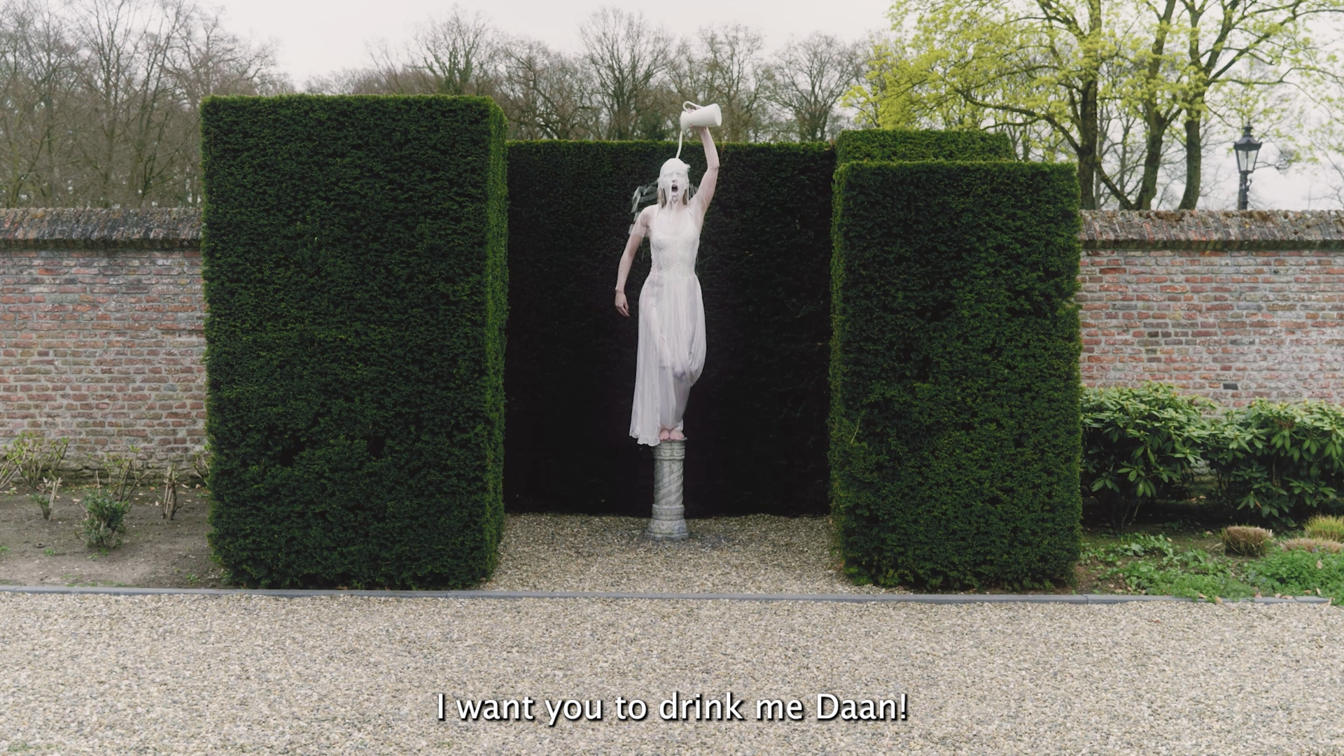 Statue of a woman with long hair, dressed in a white dress, standing on a column with her mouth open, holding a watering can above her head pouring water over her head. The statue is surrounded by tall, manicured green hedges and set against a brick wall with trees in the background. Subtitles read, "I want you to drink me Daan!"