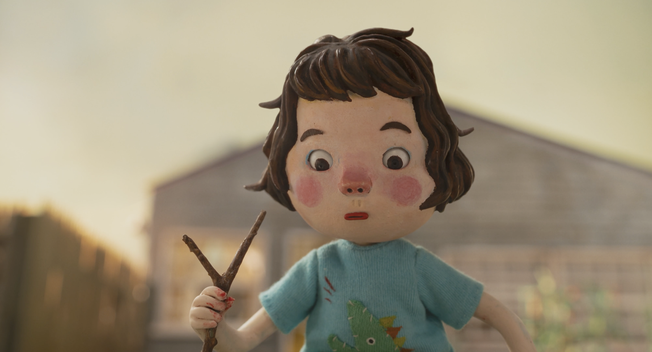 A close-up of a handmade doll with a surprised expression, brown curly hair, wearing a light blue shirt with a dinosaur design, holding a stick, set outdoors with a blurred background of buildings and a fence.