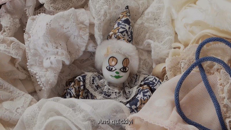 A clown doll with a white face, painted green lips, and detailed facial features, surrounded by lace and fabric, with a subtitle reading 'And quickly!'.