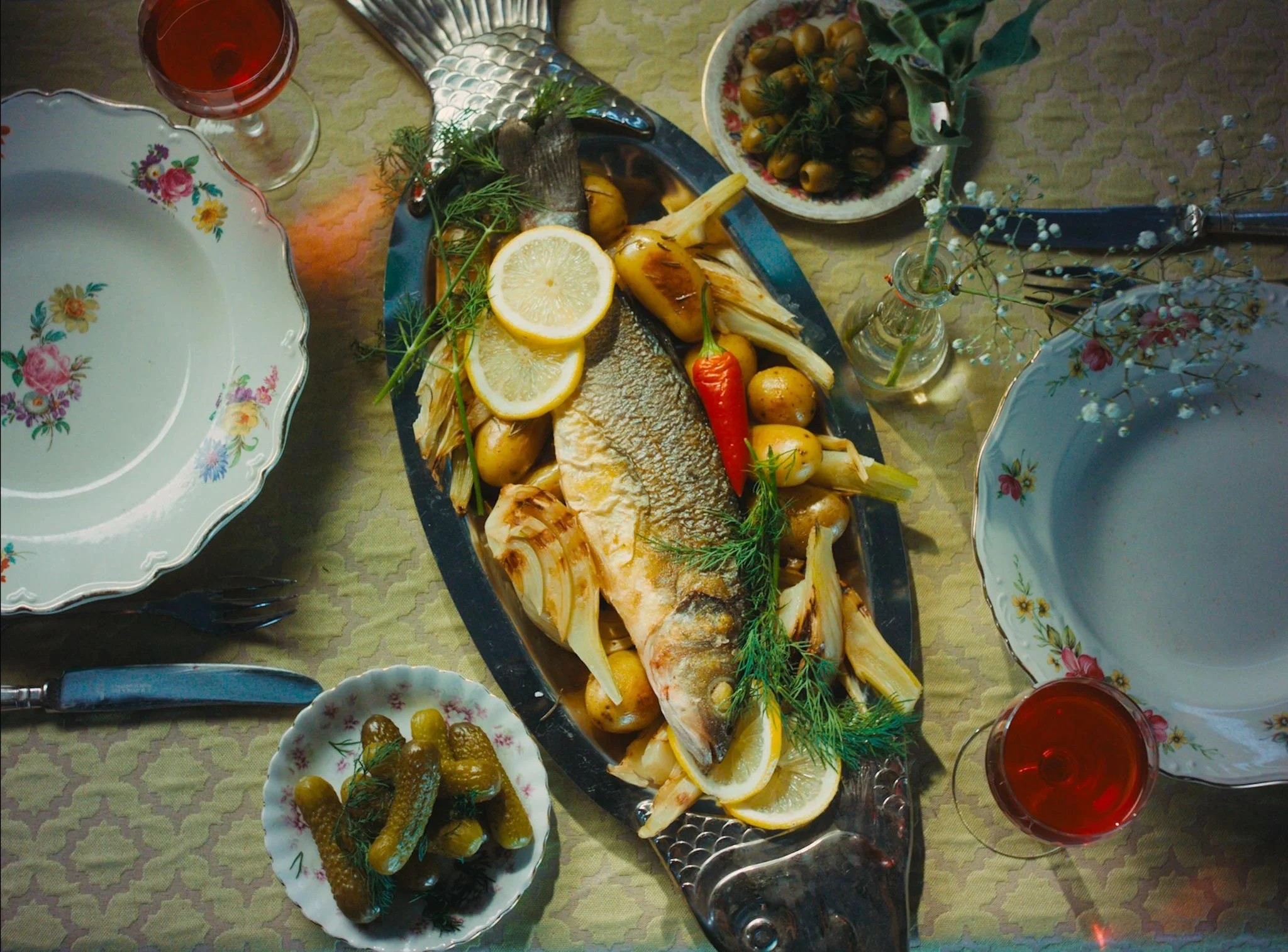 A table setting with a whole fish garnished with lemon slices, herbs, and vegetables on a black platter. There are two bowls of pickles and a small flower vase with white flowers. Two glasses of red wine are also on the table, and the tablecloth has a yellow and green pattern.
