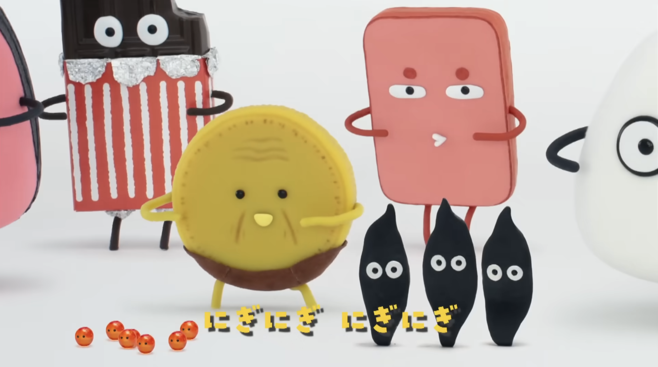 Animated characters resembling various snacks, such as a chocolate bar, popcorn box, cookie, pink rectangle, and yellow circle, with small orange balls scattered in front.