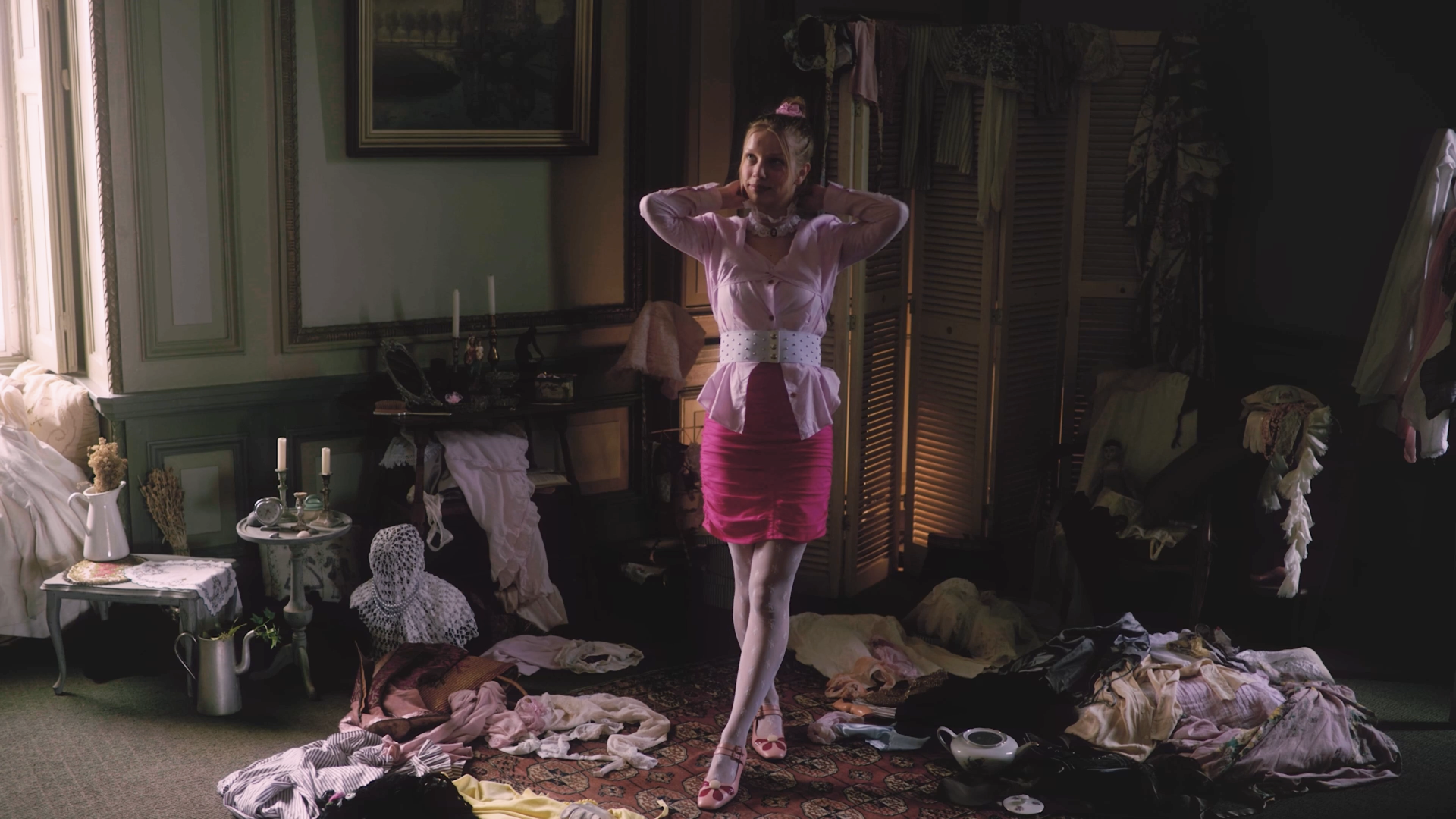 A young woman in a pink blouse, pink skirt, and white tights standing in a cluttered room with clothes and items scattered on the floor. She has her hands behind her head and appears to be adjusting her hair. There are various household objects and laundry hanging in the background.
