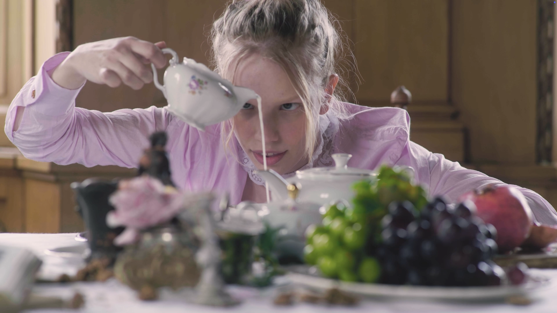 A woman with blond hair in a bun, dressed in a pink shirt, is pouring tea from a floral teapot into a cup, surrounded by various fruits and flowers on a table, in a vintage-style room.