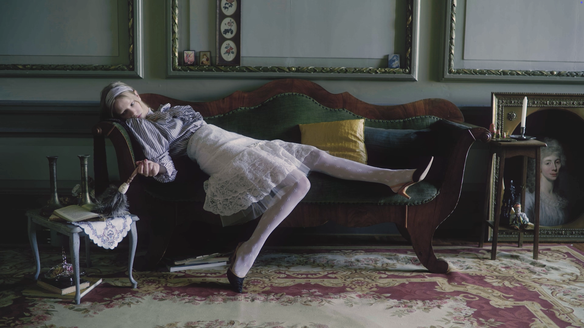 A girl lying on a vintage green velvet sofa in a decorated room with framed artwork on the wall, a side table with books and vases, and a display shelf with a portrait and objects. The girl is dressed in vintage clothing, stockings, and high heels, with a doll in her hand.