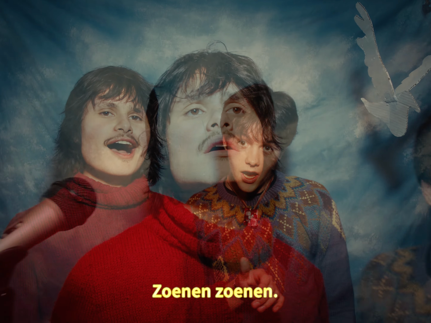A layered image of two children with questioning and surprised expressions, superimposed with a distant face of a man, with a background of a cloudy sky and a flying butterfly. Subtitles read "Zoenen zoenen."
