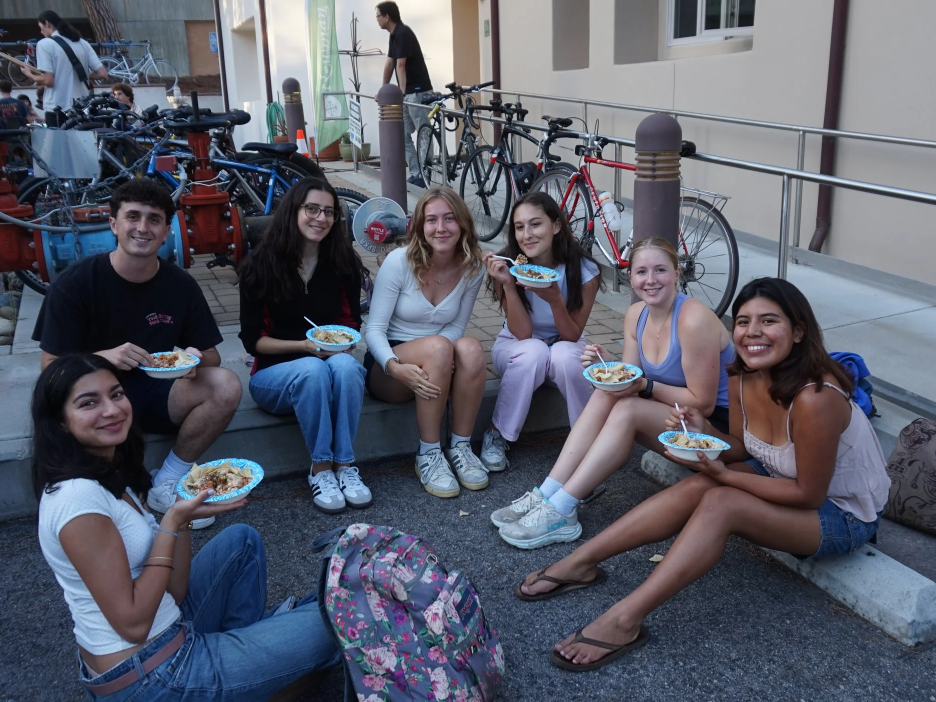 A group of seven teenagers sitting and kneeling on the ground, holding bowls of food with spoons, smiling at the camera. They are outdoors near a building, with bicycles and biking gear in the background.