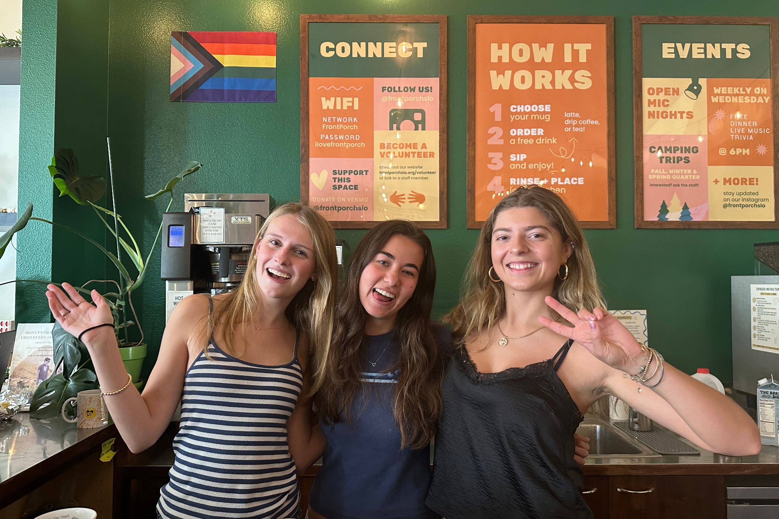 Three young women smiling and posing in a kitchen with colorful signs on a green wall behind them. The signs display information about WiFi, how it works, events, and support options.