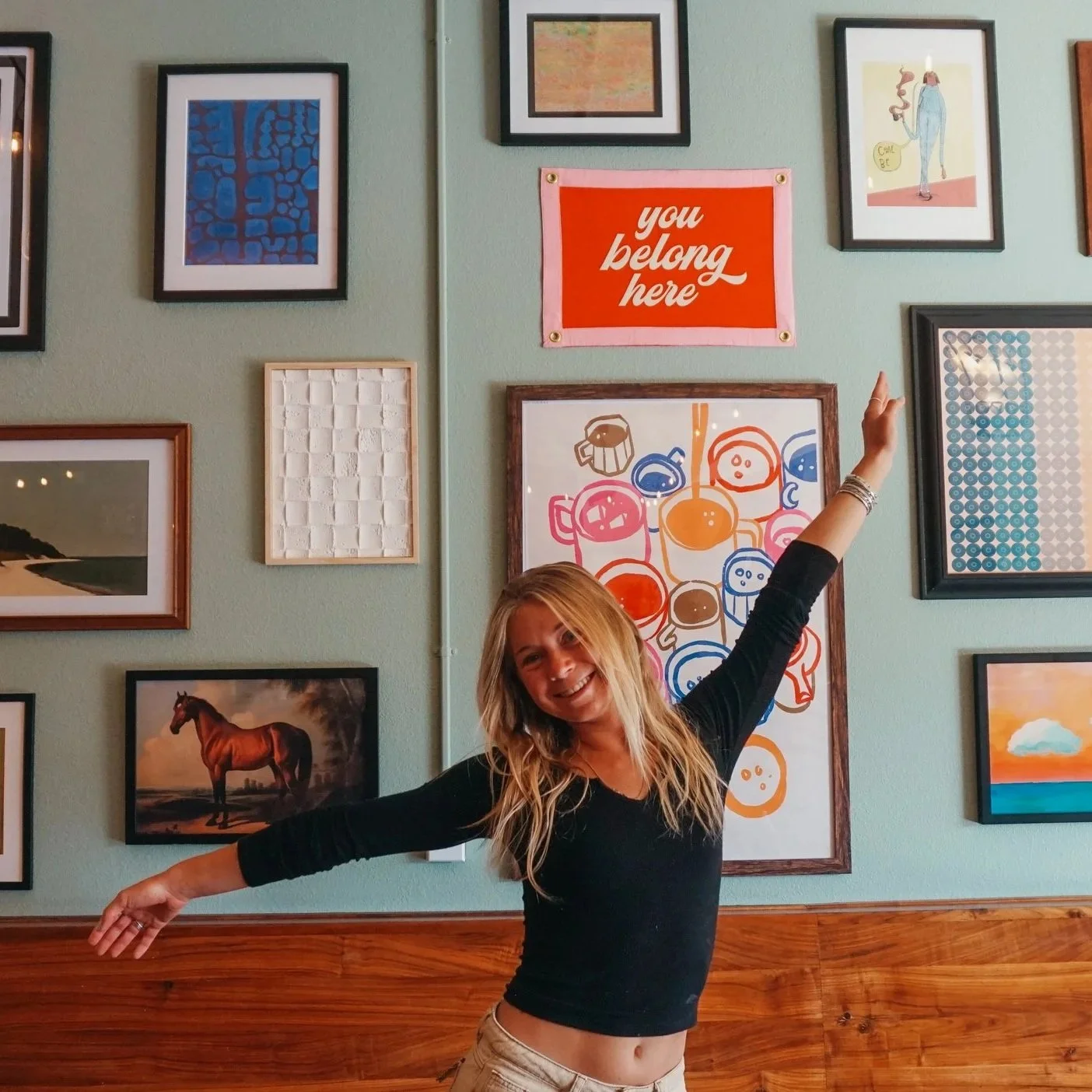 A woman with long blonde hair smiling with arms outstretched standing in front of a gallery wall with various framed artworks including a horse, abstract art, and a sign that says 'you belong here'.