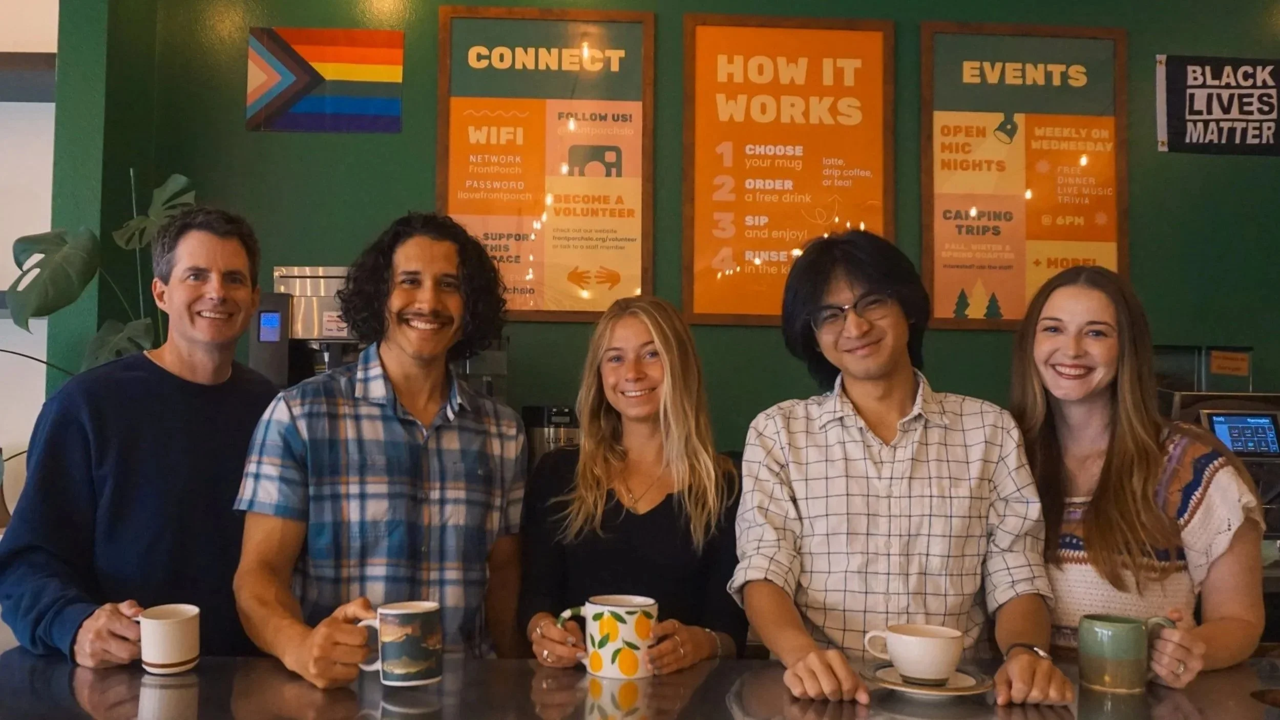 Group of five people smiling and holding mugs in a cafe or coffee shop with green walls, framed posters including a rainbow pride flag, and a plant in the background.