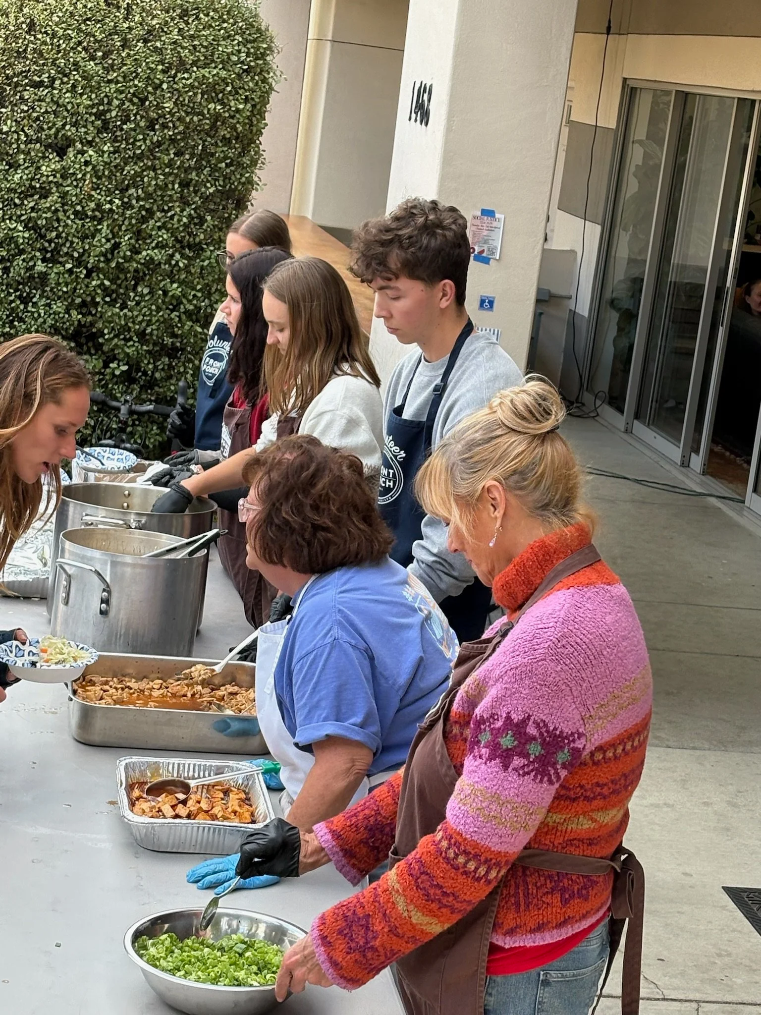 People serving themselves food at a buffet table outdoors, including salads and cooked dishes.