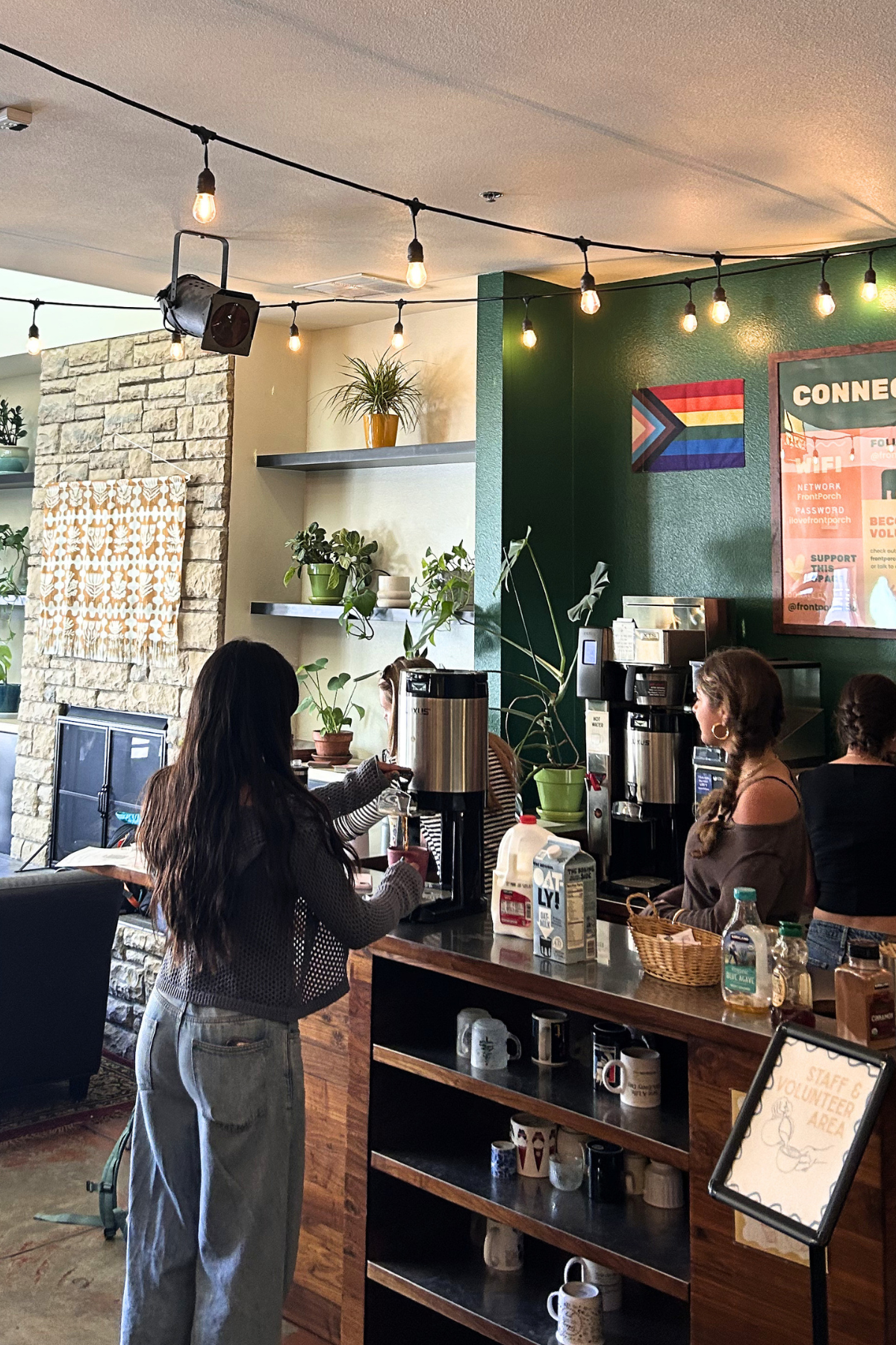 Inside a coffee shop with three women standing at the counter, various coffee-making equipment, and decorative plants on shelves. String lights hang from the ceiling, and there is a stone wall with artwork and a green-painted wall with a Wi-Fi sign.