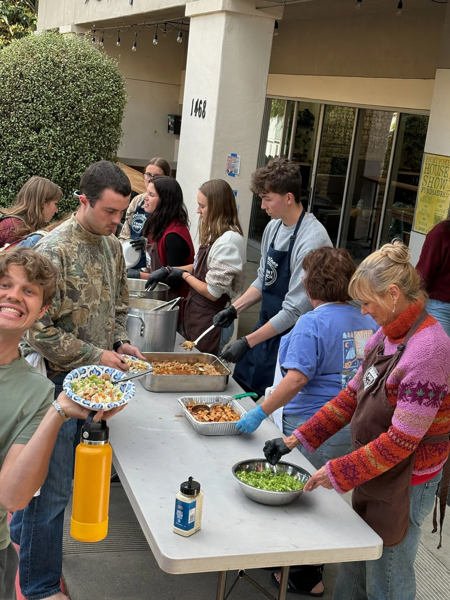 People serving themselves food at a buffet table outdoors during a gathering. There is a man in the foreground holding a plate of food, and several others are behind the table selecting dishes. Various food items and bowls are set out on the table.