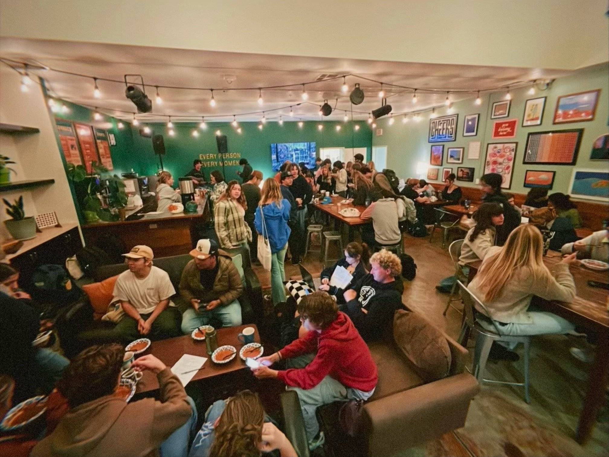 A crowded indoor space with people socializing and sitting at tables. The walls are decorated with framed artwork, and string lights hang from the ceiling, creating a cozy atmosphere.