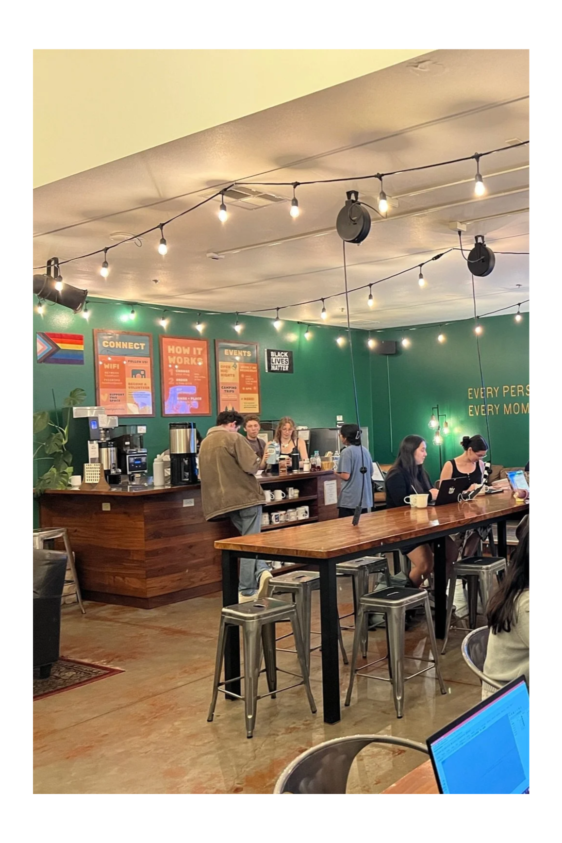 A busy coffee shop with several customers at the bar and tables, string lights hanging from the ceiling, and green walls decorated with posters and a black Lives Matter sign.