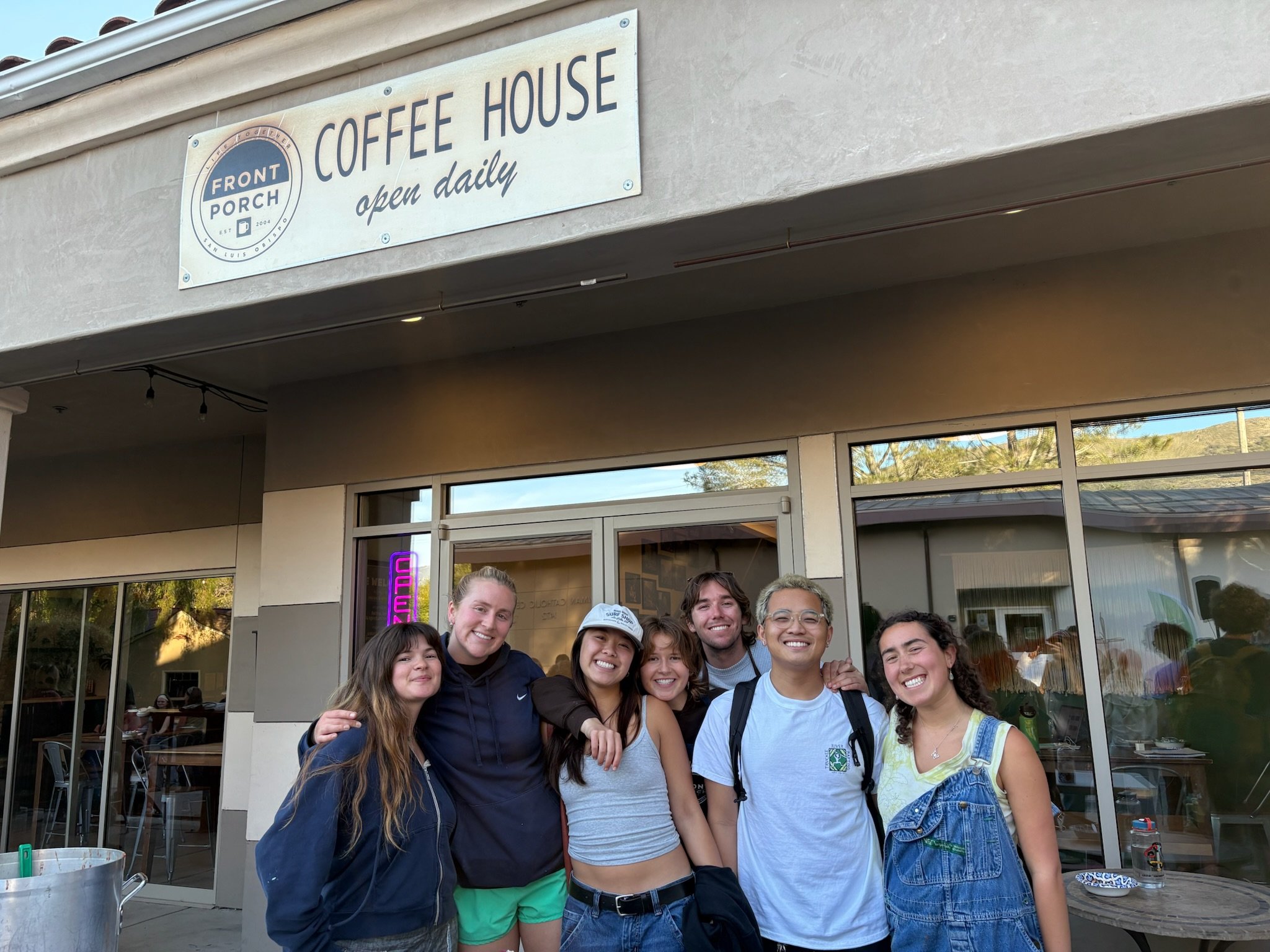A group of eight smiling young people standing together outside a cafe, with a sign that reads 'Coffee House open daily,' in front of large windows reflecting trees and a blue sky.