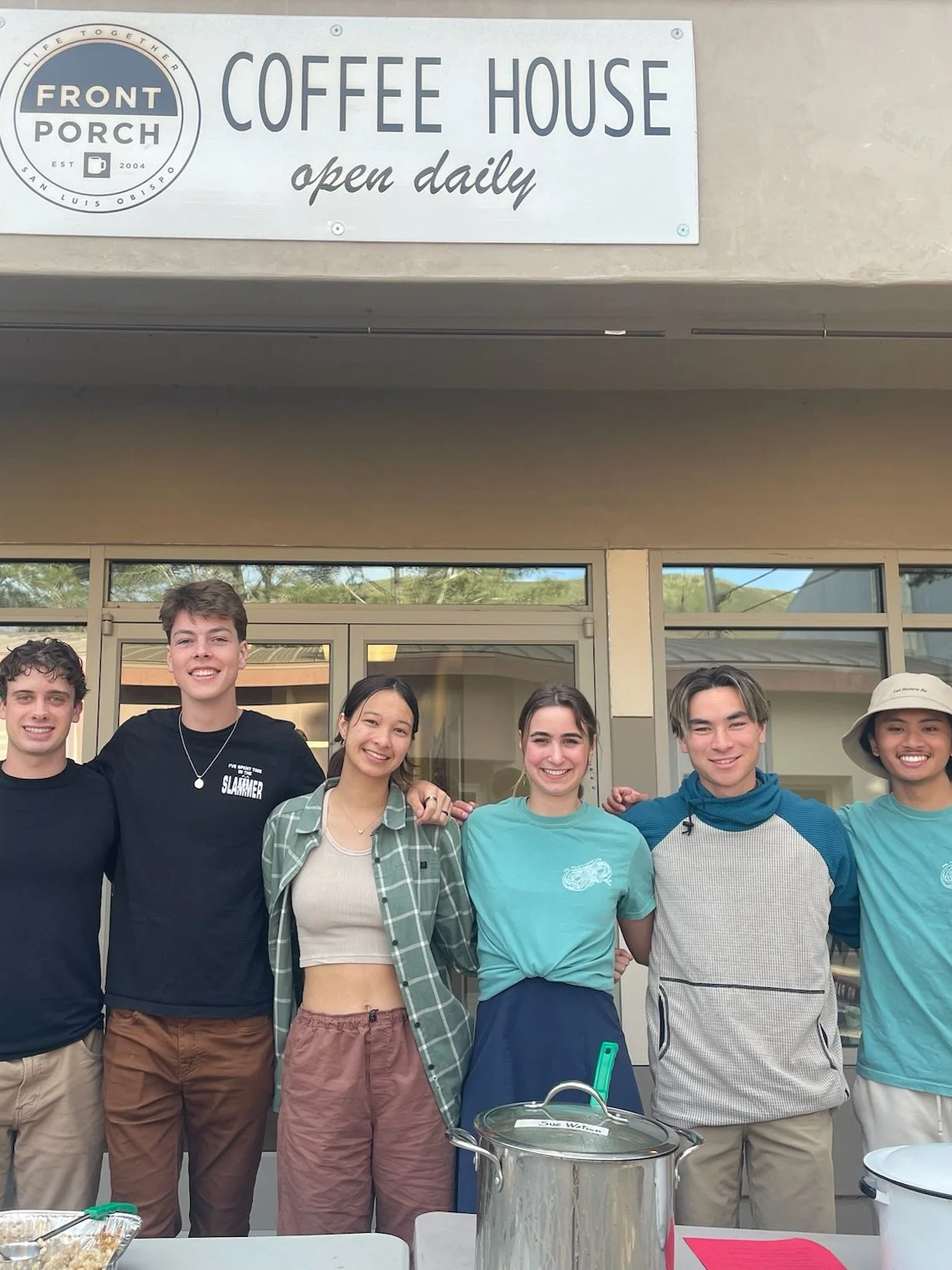 Group of six young people smiling and standing together outdoors in front of a coffee shop with a sign that reads 'Coffee House, open daily.'