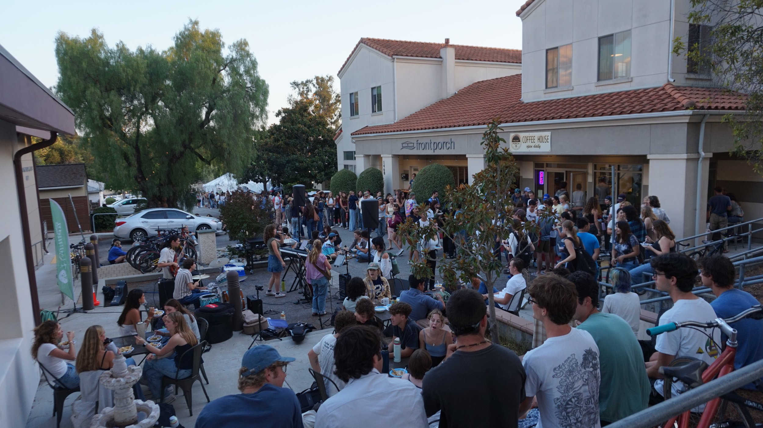 Crowd of people gathered outside a building with a sign that reads 'frontporch' and 'coffee house' during late afternoon.