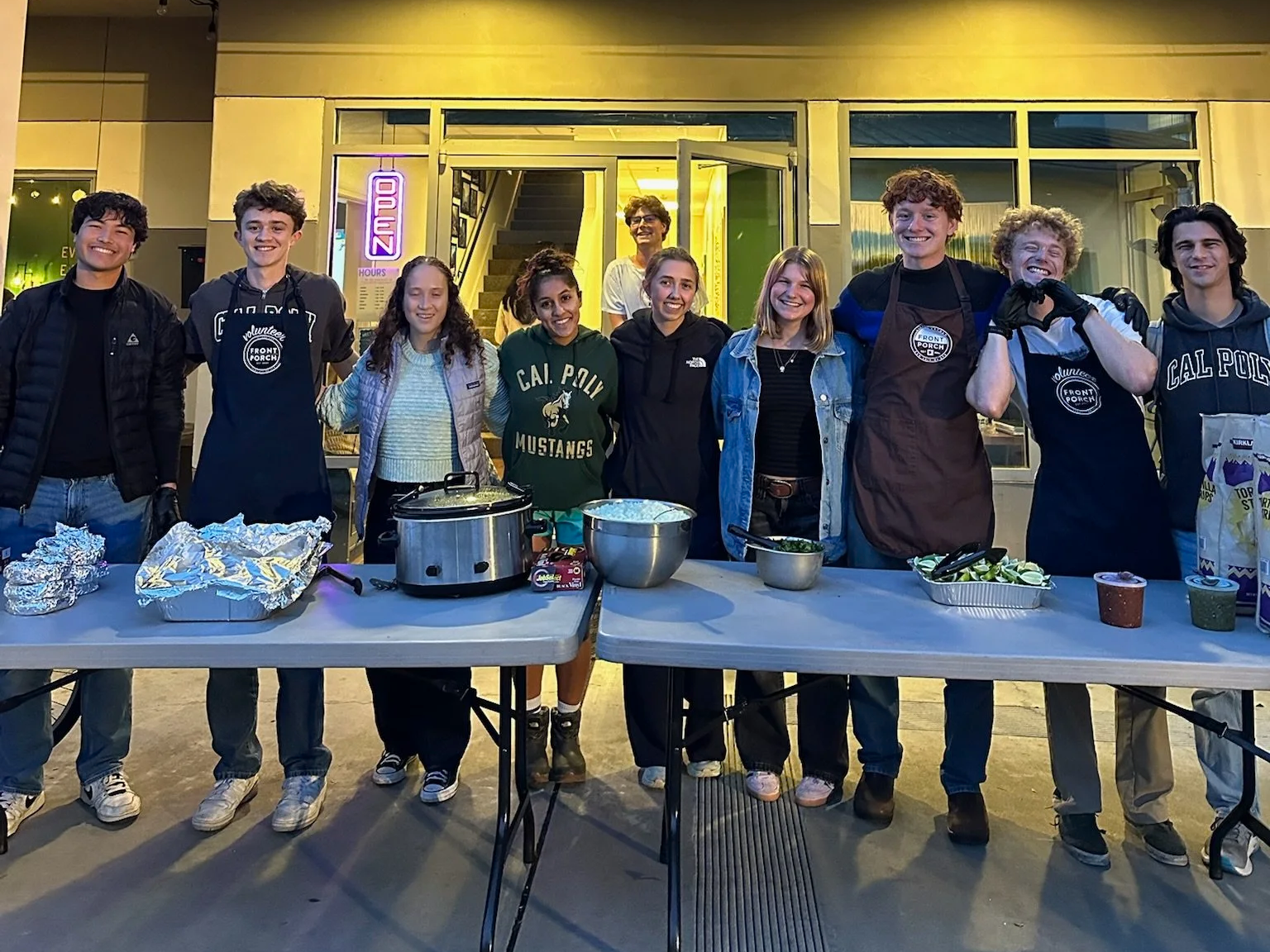 A group of ten young people standing behind a table with food at an outdoor event, smiling for a photo at night.