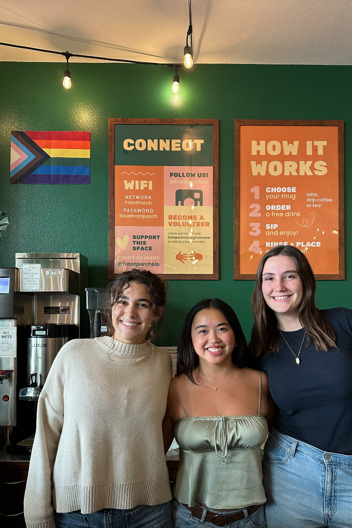 Three women standing together in a coffee shop, smiling at the camera, with a green wall and informational posters behind them.