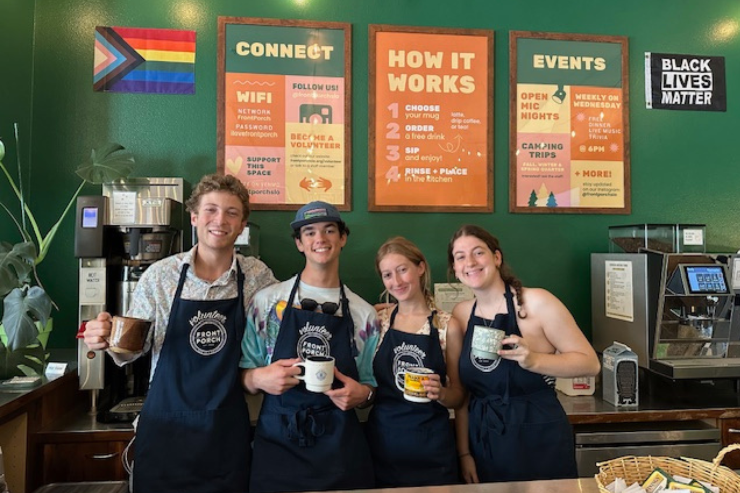 A group of four young people smiling and holding cups of coffee while standing behind a counter, wearing aprons in a coffee shop. The background features colorful signs about WiFi, event details, and how the shop works, with a green wall and various equipment.