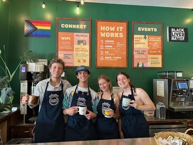 Four young people working at a coffee shop, smiling behind the counter, wearing black aprons with a logo, in a green-walled cafe with signs about events and how it works.