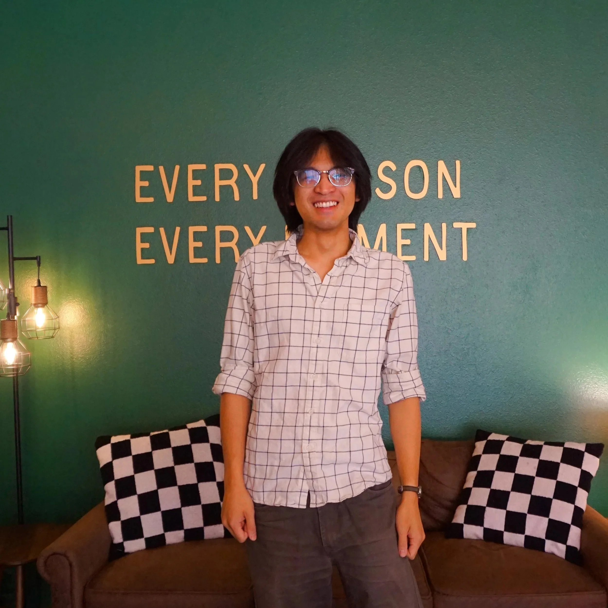 A man with glasses smiling, standing in front of a green wall that has the words 'EVERY PERSON EVERY MOMENT' written on it. There is a checkered-pattern cloth or backdrop partially visible in front of him.