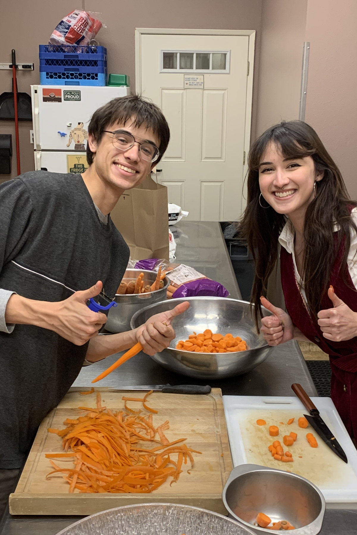 Two people smiling and giving thumbs up while preparing chopped carrots in a kitchen. There is a cutting board with shredded carrots, a knife, and bowls of chopped carrots on the counter.