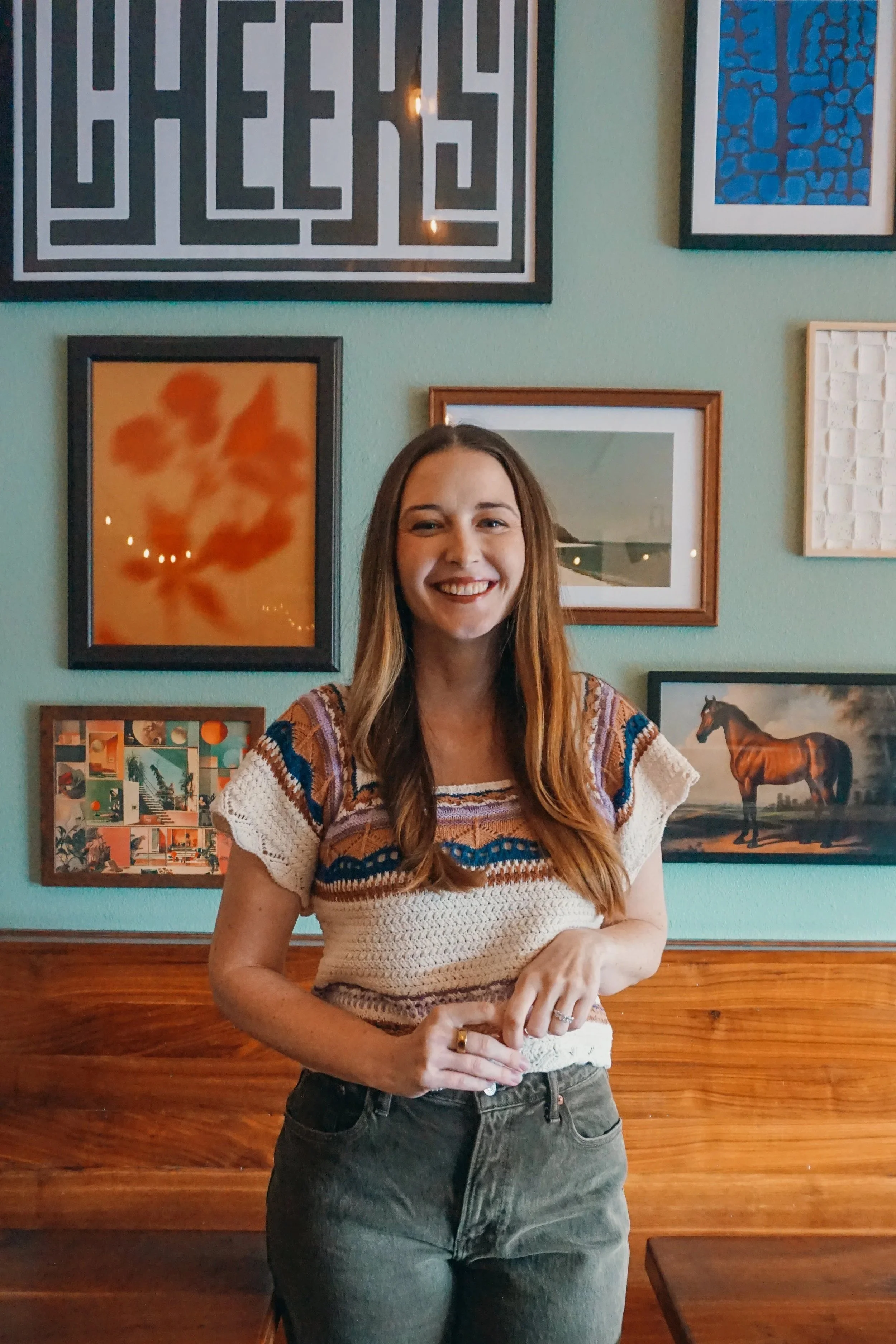 A woman with long light brown hair smiling, standing in front of a gallery wall with various framed artworks, including an abstract piece, a photo of a horse, and a colorful collage, in a cozy room.
