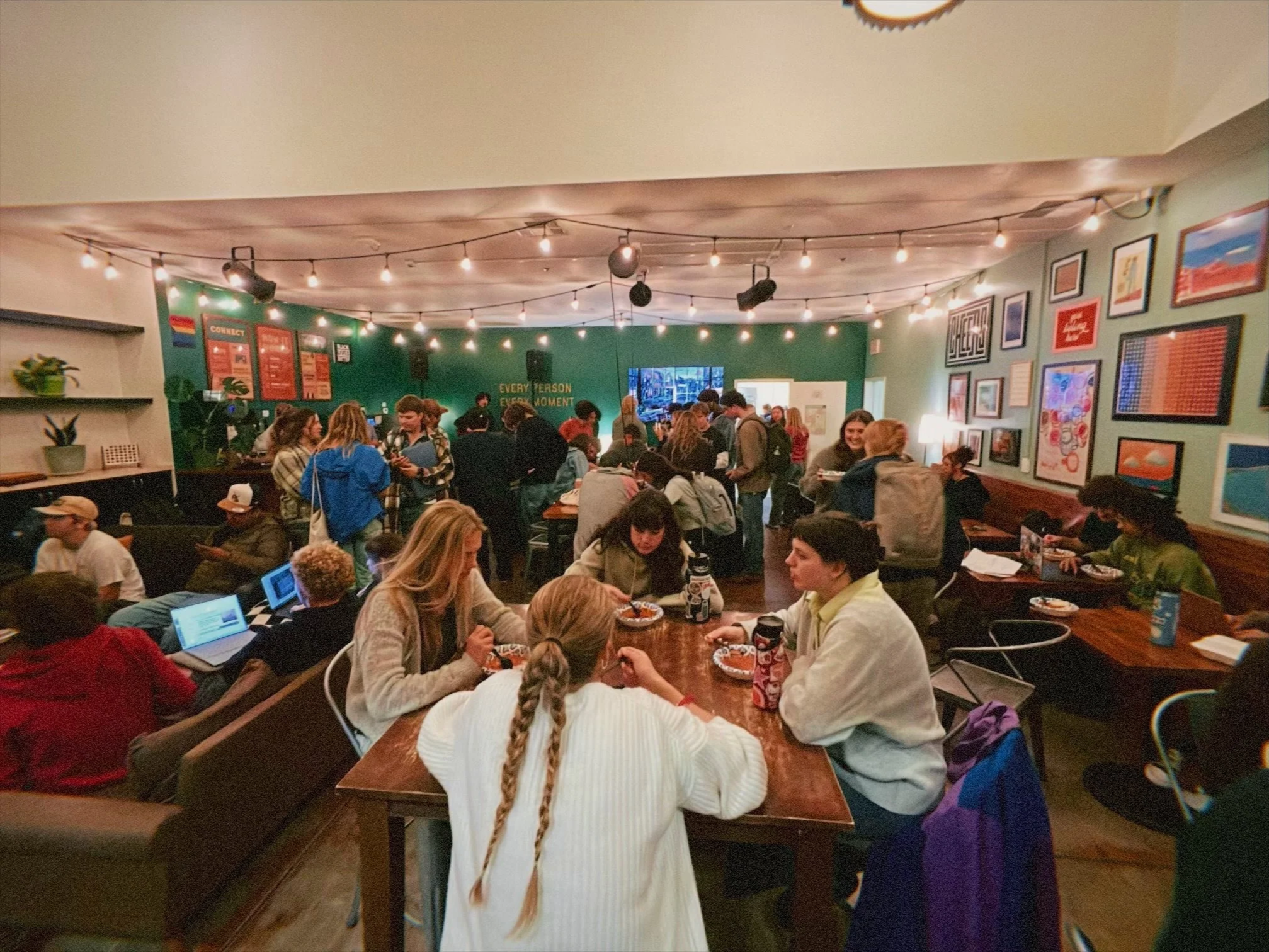 A crowded indoor gathering with people eating at a long table, some using laptops, in a room decorated with string lights, framed artwork, and a green wall with the quote 'Every person is an opportunity'.