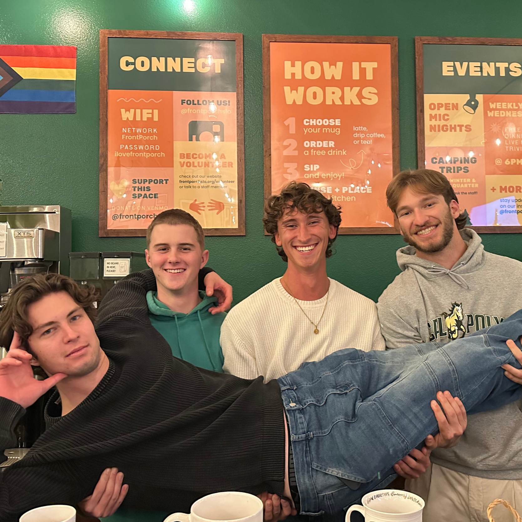 Four young men smiling and posing together, with one man holding another horizontally in front of a green wall with informational posters, behind a table with white mugs.