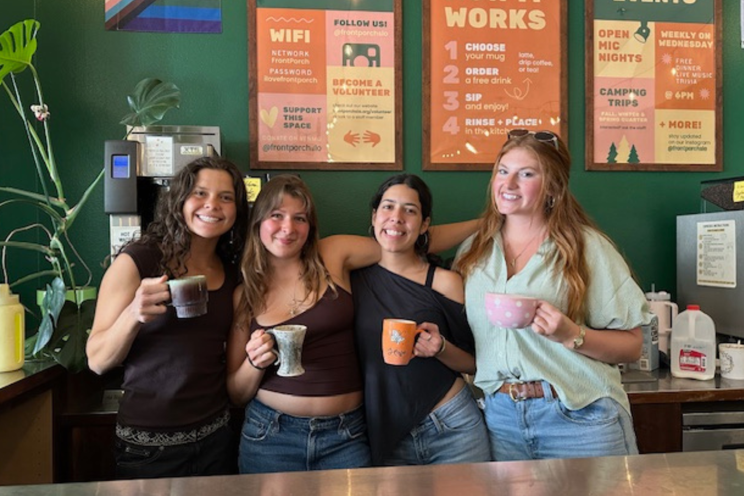 Four women smiling and holding coffee mugs in a coffee shop with green walls and various informational signs behind them.