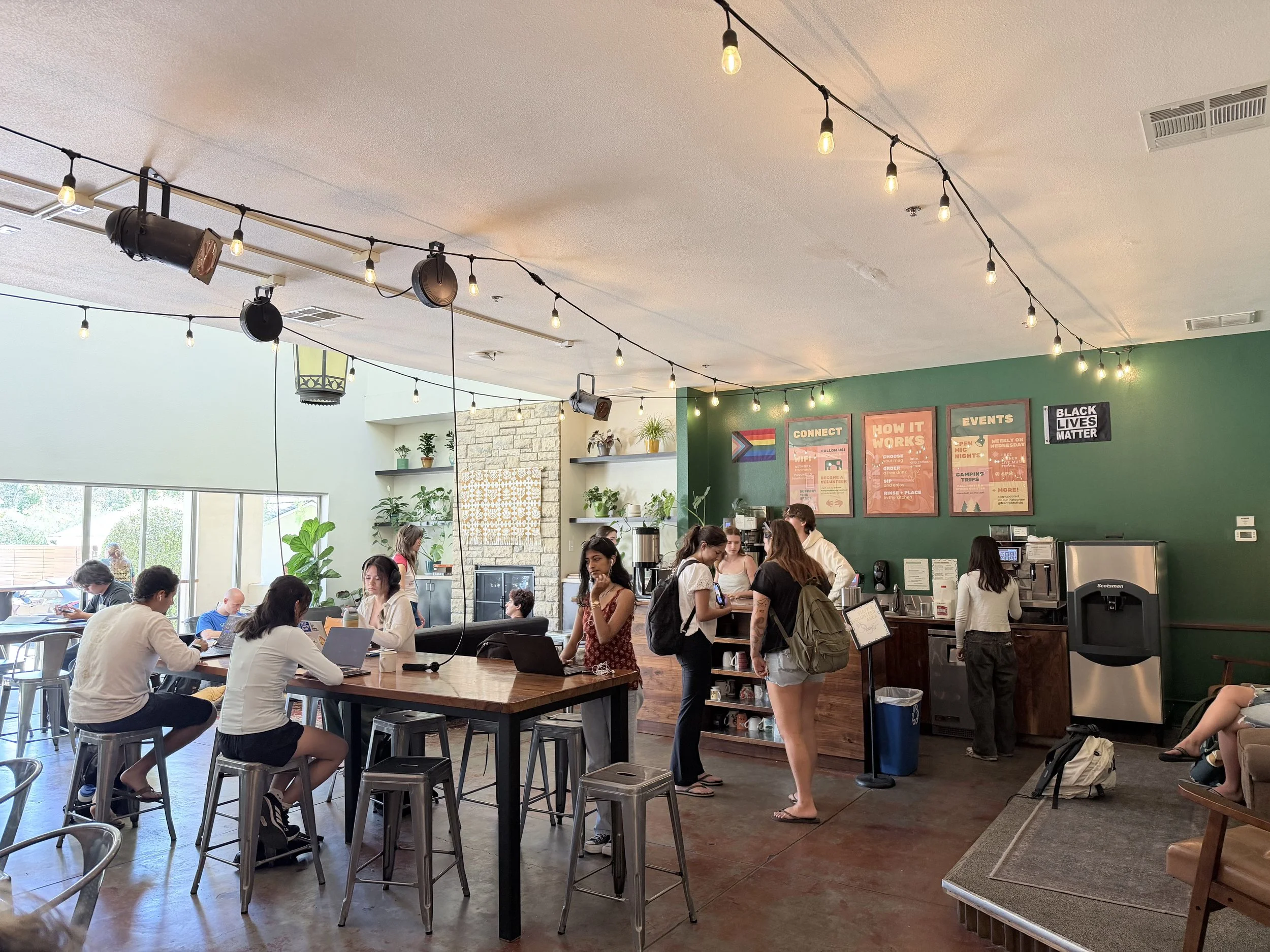 People are gathered inside a bustling cafe or coffee shop, some sitting at tables working on laptops and others standing in line or ordering at the counter. The space features string lights hanging from the ceiling, a green accent wall with posters, and large windows letting in natural light.