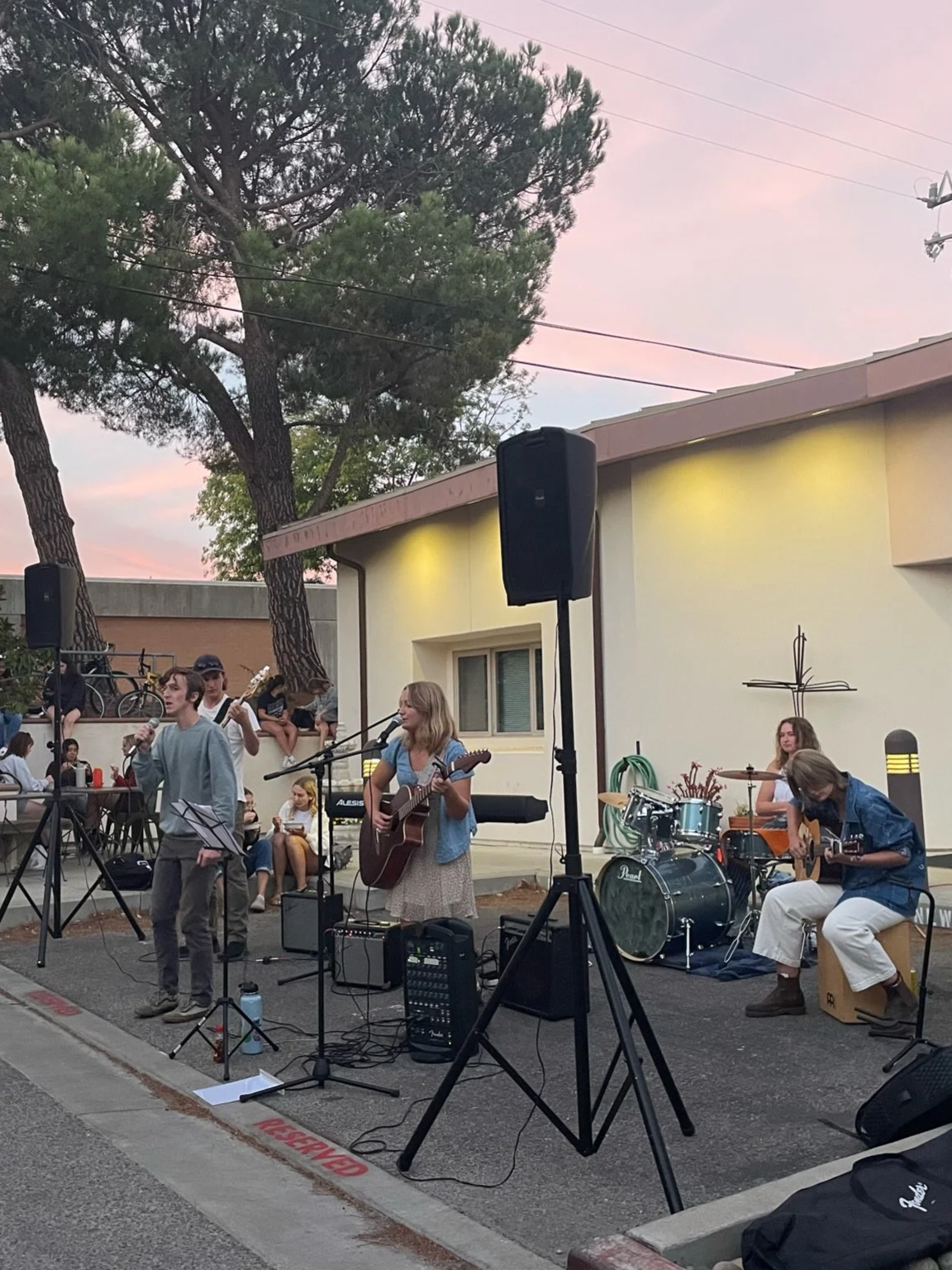A group of young musicians performing outdoors in a parking lot at sunset, with trees and a building in the background.
