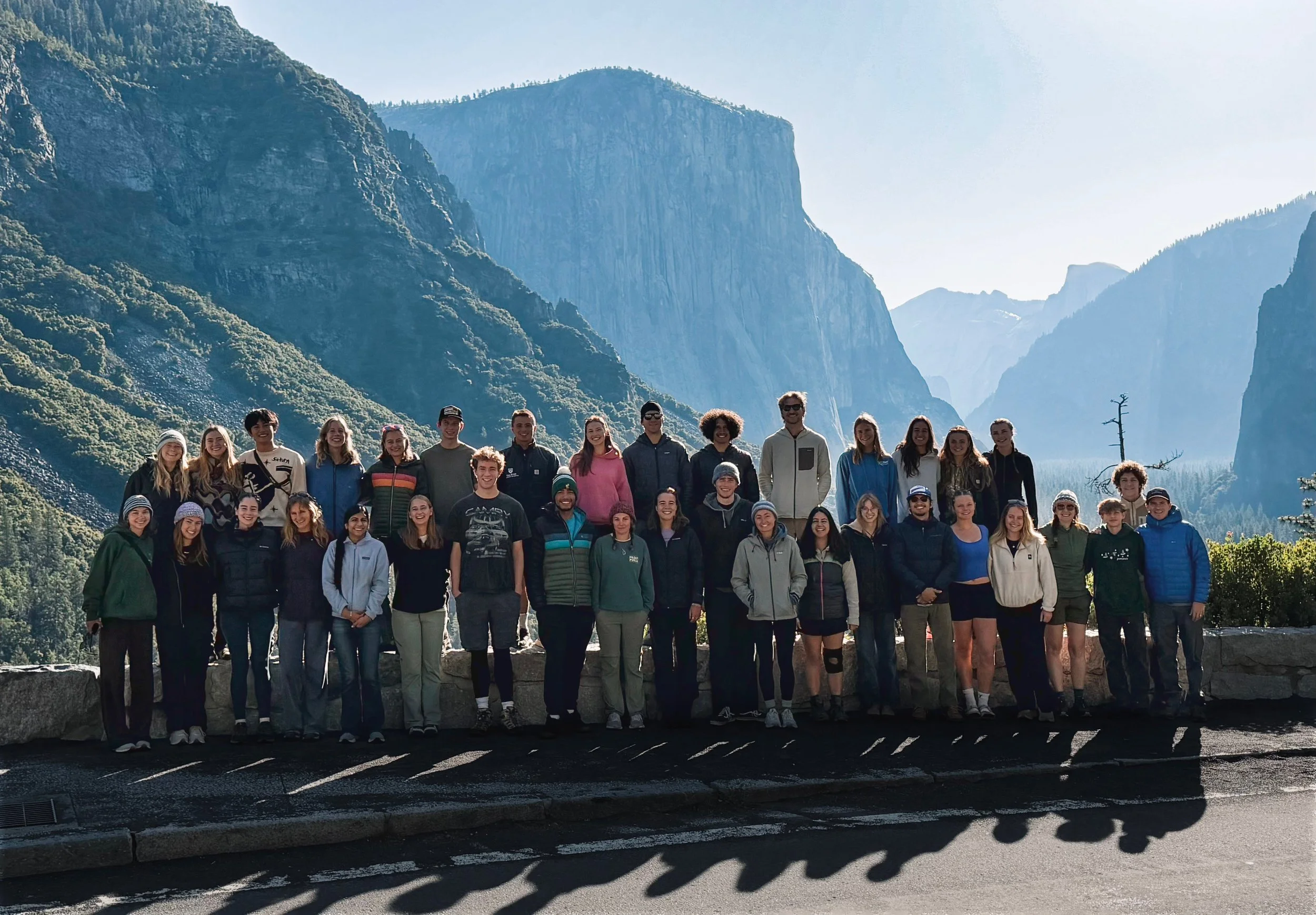 A large group of people standing outdoors in front of mountainous scenery, smiling for a group photo.