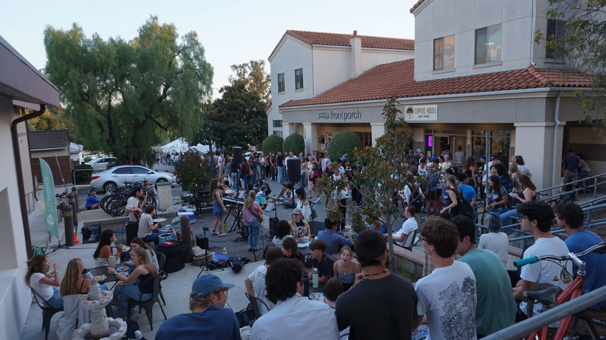 Crowd gathering outside a building with a sign that reads 'front porch' and a coffee shop sign, during early evening, with people sitting and standing, some playing music and others socializing.